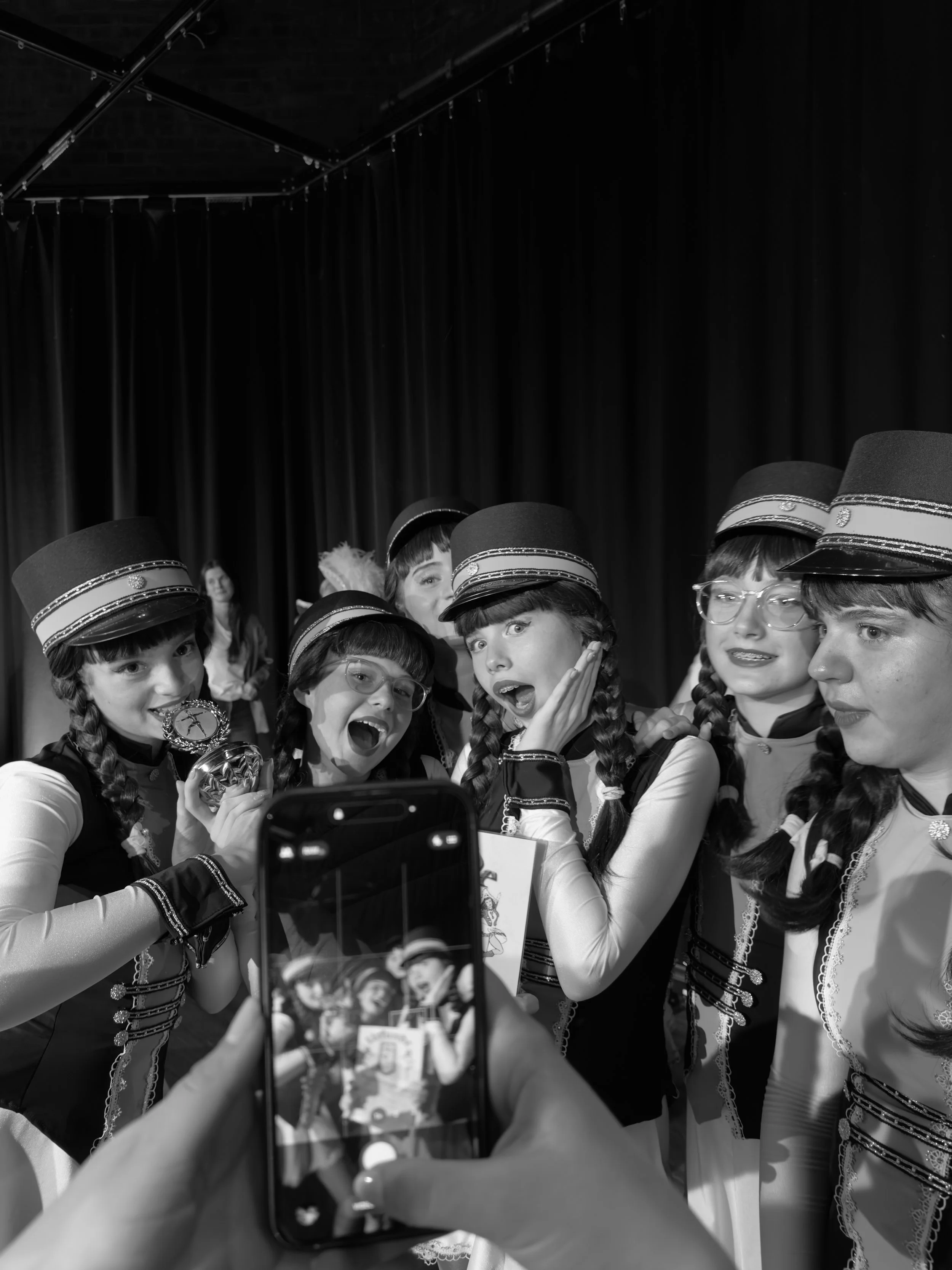 Junge Mädchen in traditionellen Kostümen und Hüten posieren für ein Foto auf einer Bühne mit schwarzem Vorhang im Hintergrund. Eine Person hält ein Smartphone, um das Foto zu machen.