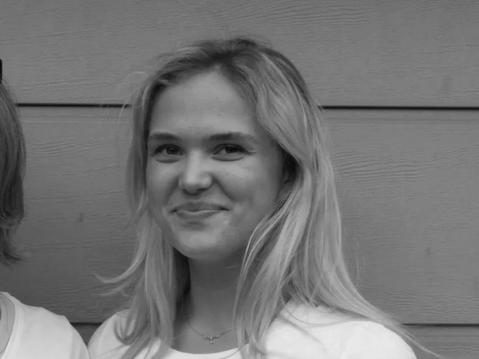 Eine lächelnde junge Frau mit mittellangen, blonden Haaren trägt ein weißes Oberteil und eine zierliche Halskette mit Anhänger. Sie steht vor einer graublauen Holzwand.