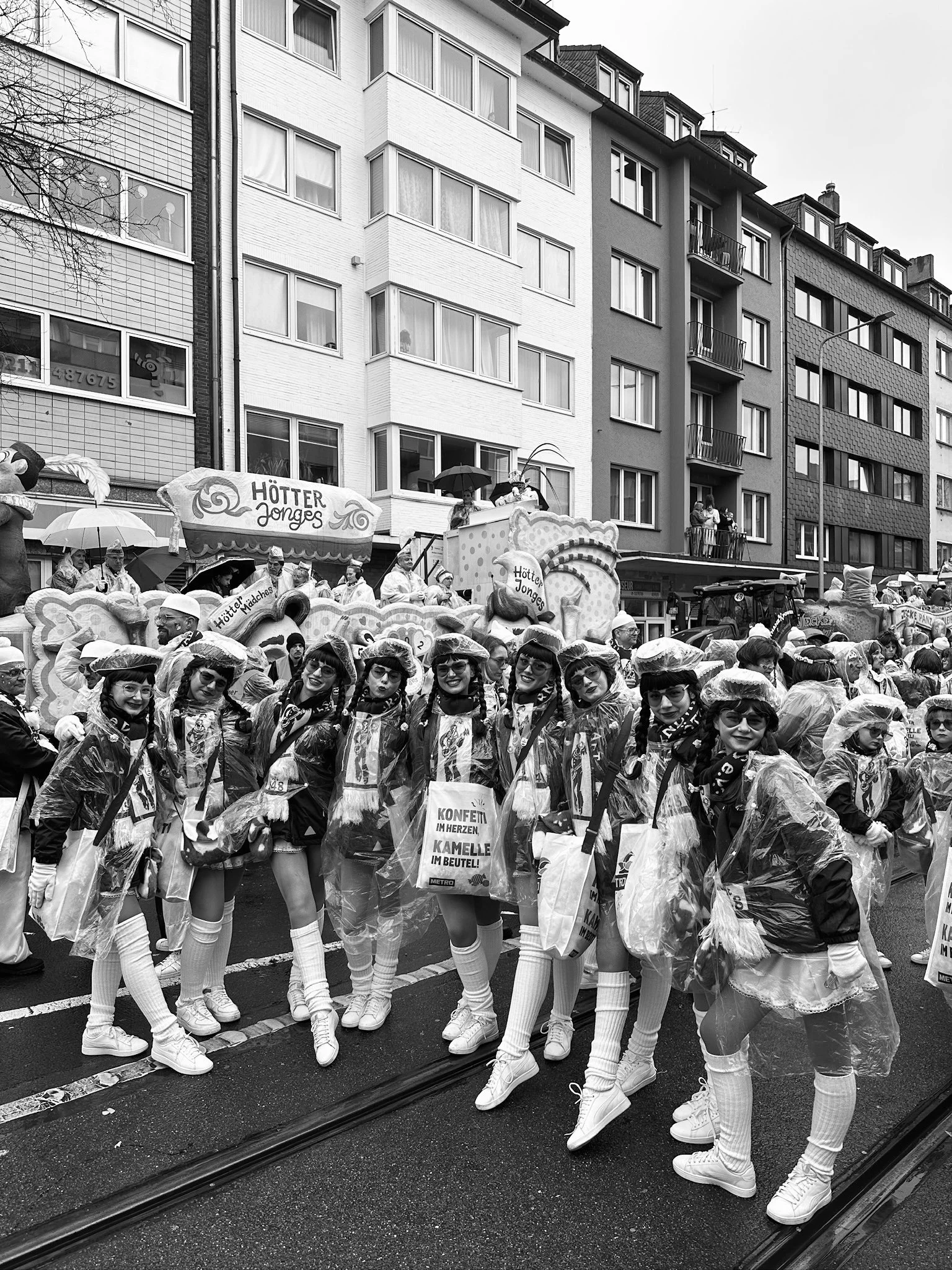 Jugendliche in Kostümen und Regenmänteln bei einem Faschingsumzug auf der Straße, im Hintergrund Häuser und ein Wagen mit einem Plakat 'Hötter Jonges'.