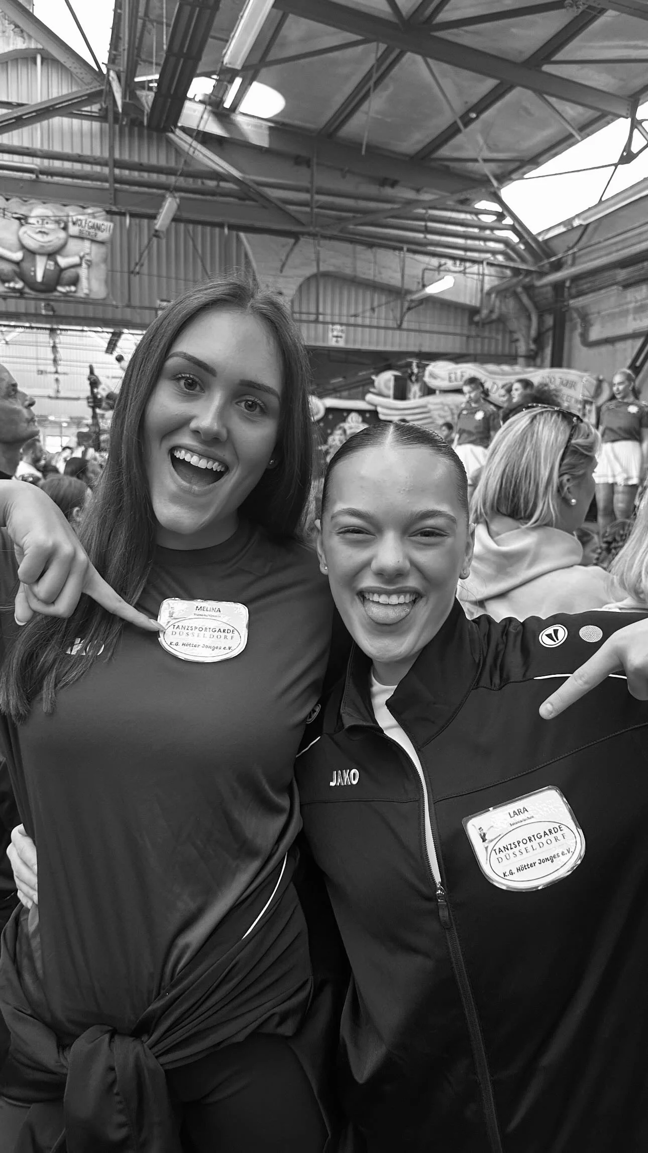 Zwei lachende junge Frauen in Sportbekleidung, die in einer Halle mit anderen Menschen stehen, einer zeigt auf ihren Namenstag-Aufnäher, im Hintergrund sehen wir andere Frauen in Sportkleidung auf einer Bühne.
