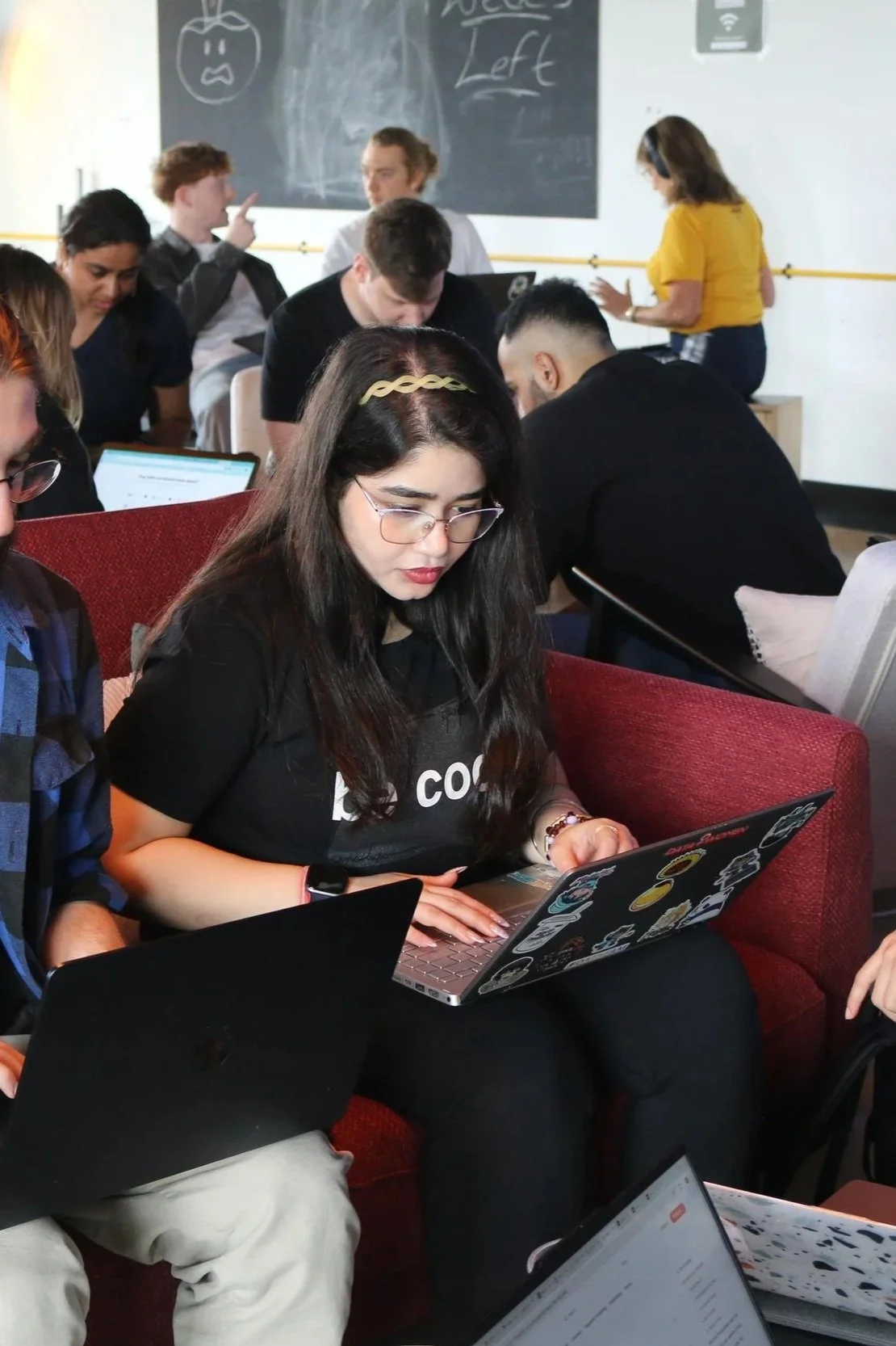 Young woman wearing glasses and a black t-shirt using a laptop with stickers, surrounded by other young adults working on laptops in a classroom or conference setting.