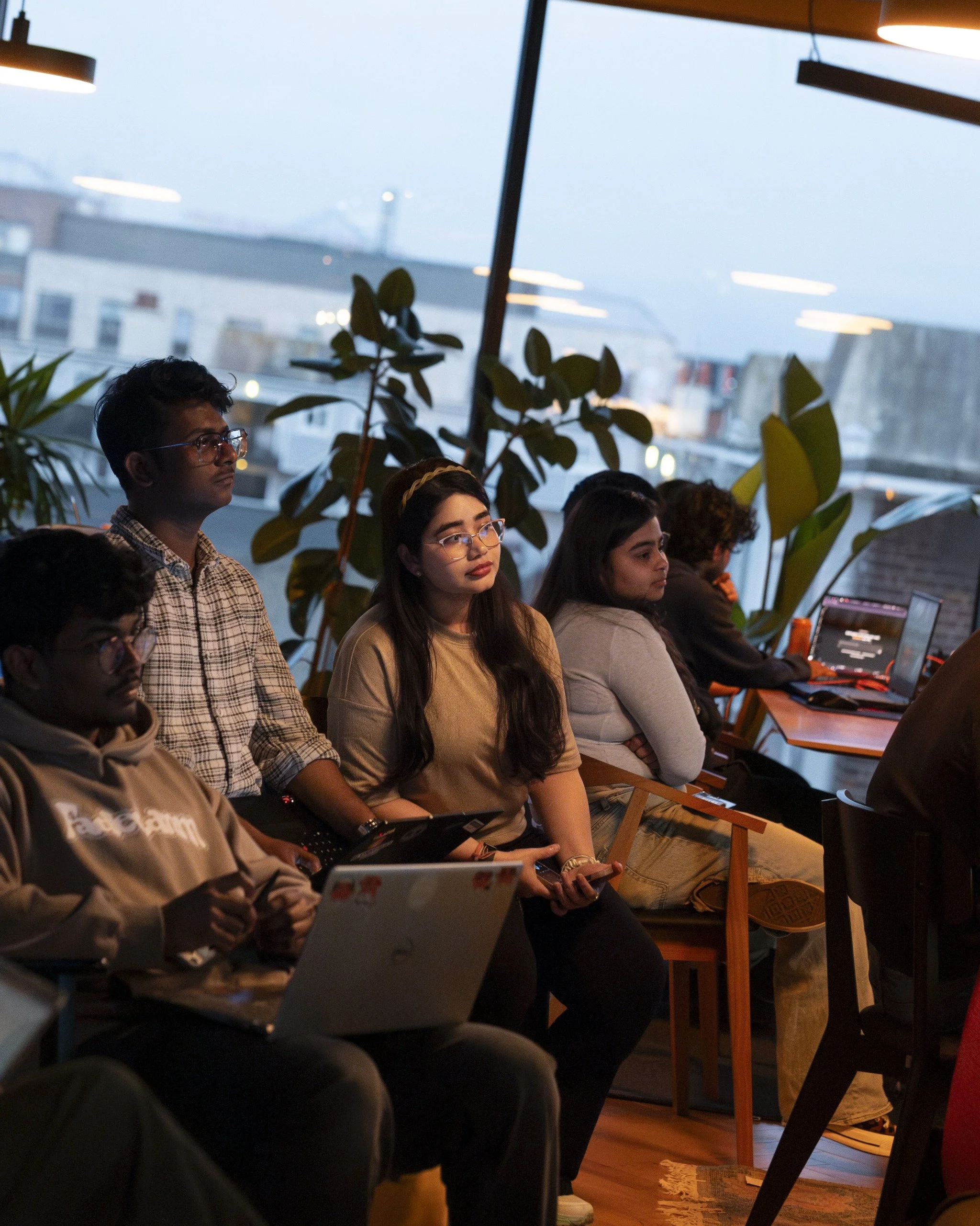 People sitting in a dimly lit room, watching a presentation or screen, with laptops and notebooks in front of them. Large windows with a city view and some indoor plants are visible.