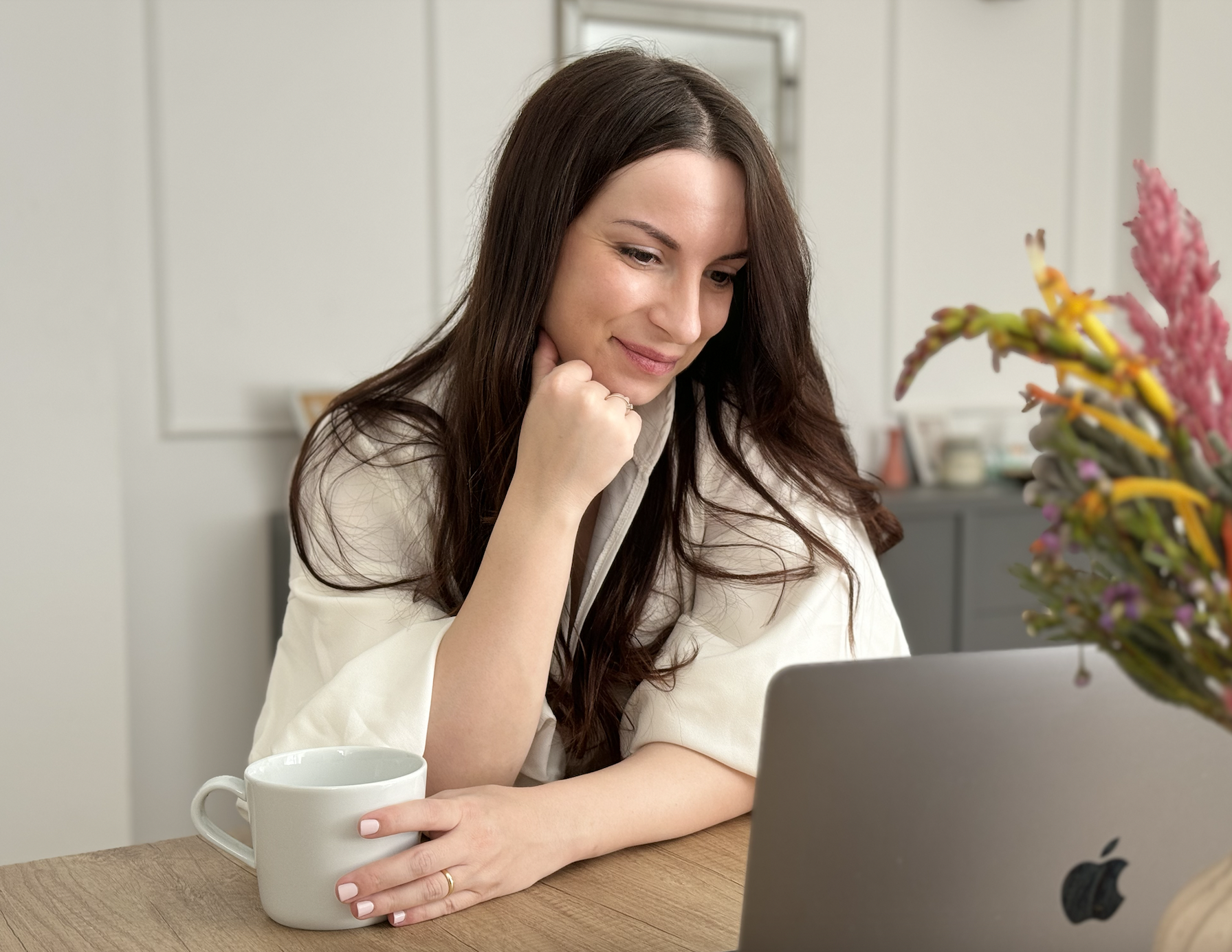A woman with long brown hair sitting at a table, looking at a laptop. She is holding a coffee mug in her left hand, resting her chin on her right hand, smiling while looking at the screen. There are colorful flowers in a vase on the table.