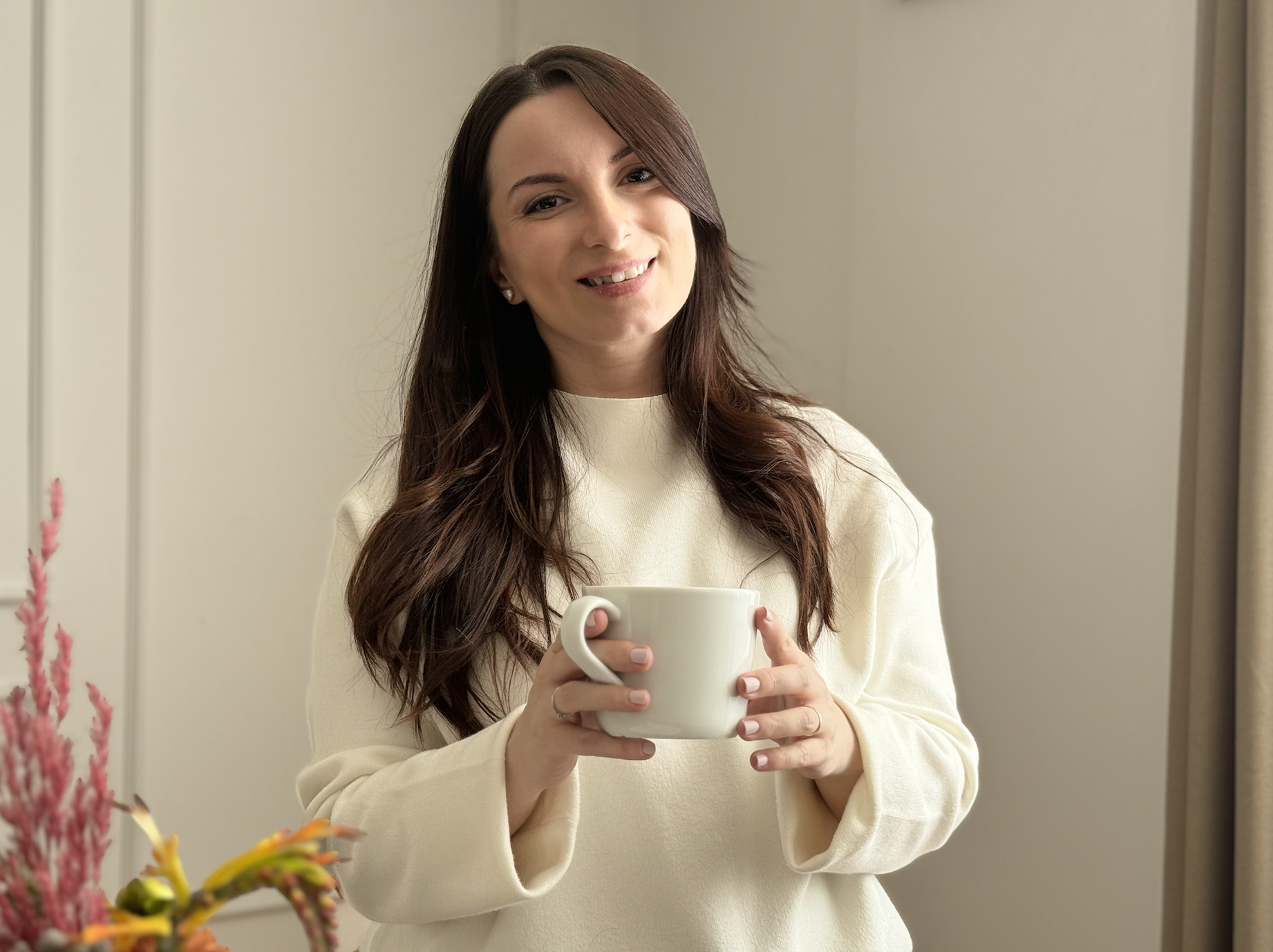 A smiling young woman with long brown hair, wearing a cream-colored sweater, holding a white mug with both hands, standing indoors near a beige wall and a curtain.