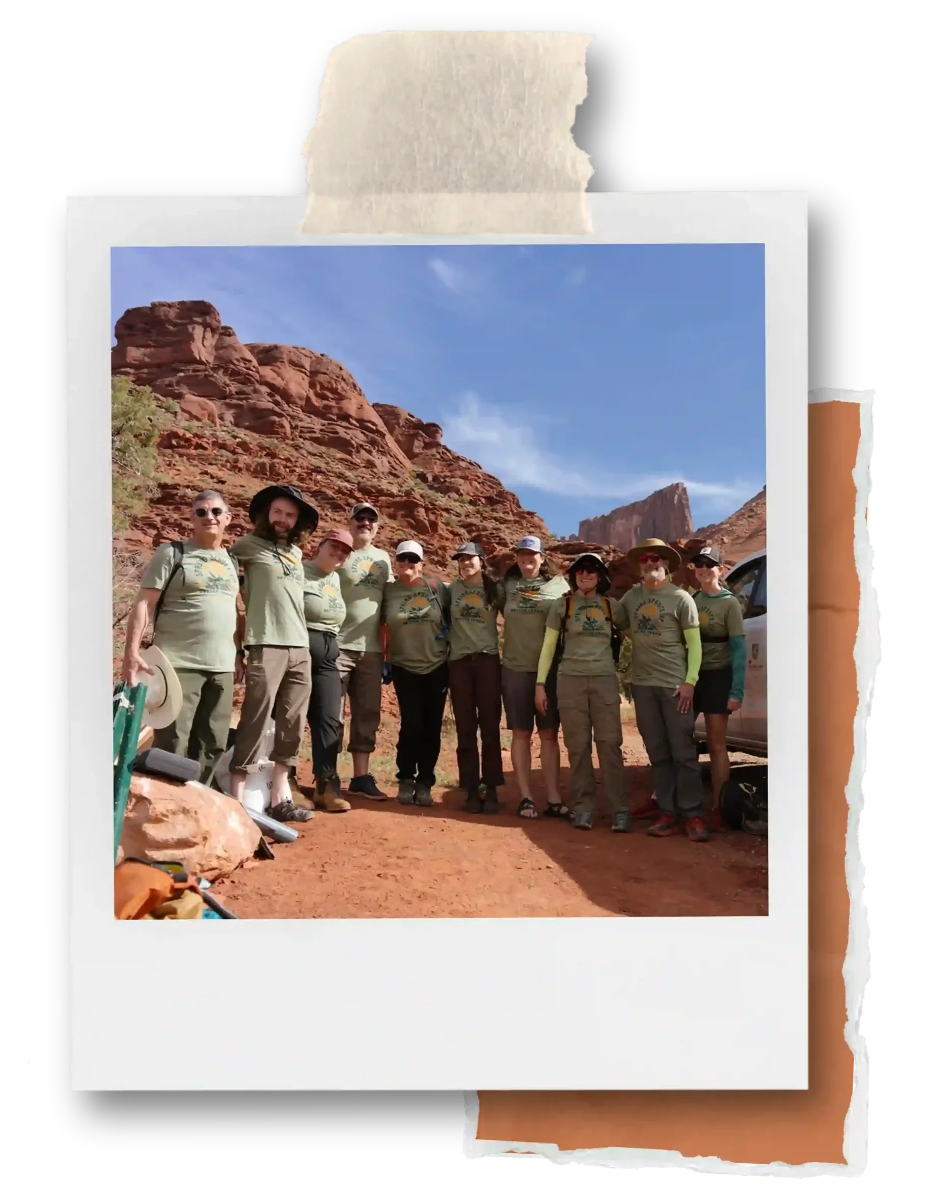 A group of people wearing outdoor clothing and hats standing in a desert landscape with red rock formations under a blue sky.