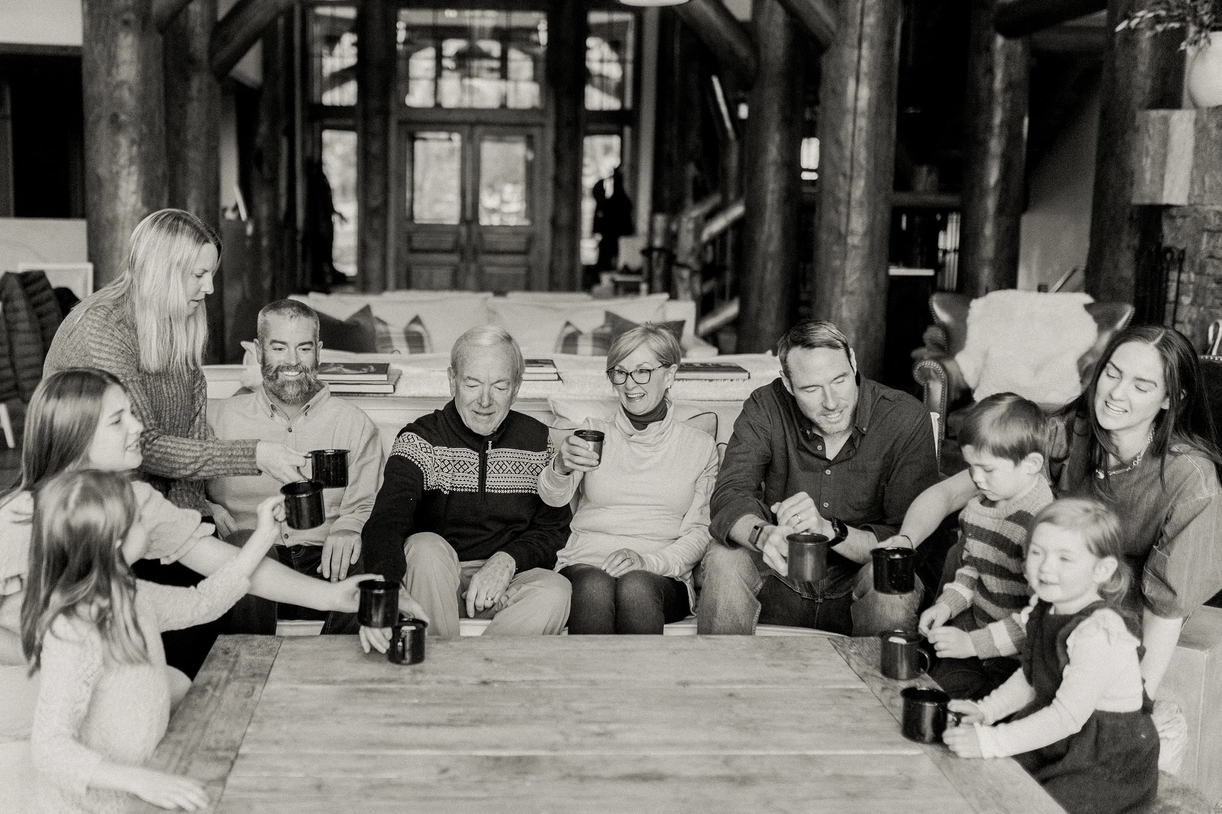 Family gathering in a cozy living room, sitting on a couch and around a table, smiling and holding coffee mugs.
