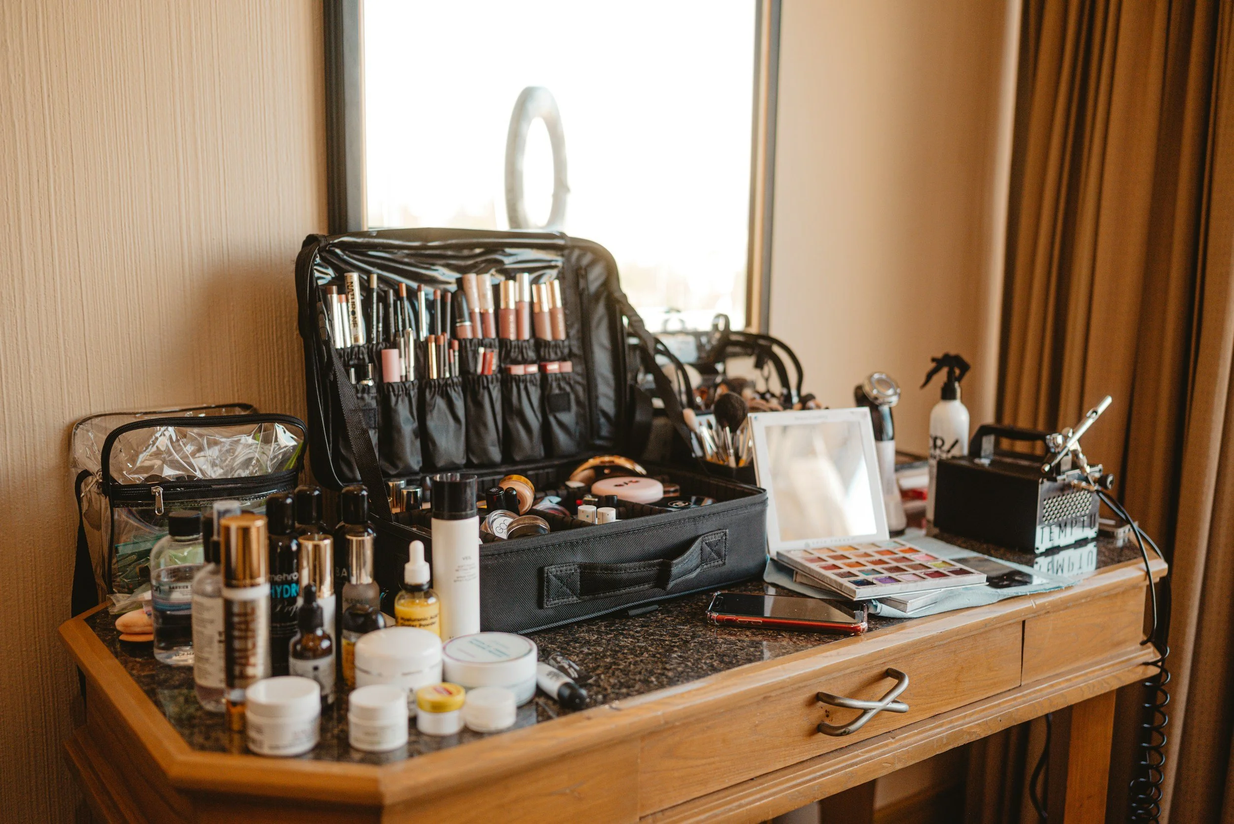 A wooden table near a window with various makeup and skincare items, including brushes, bottles, jars, a mirror, and a makeup palette.