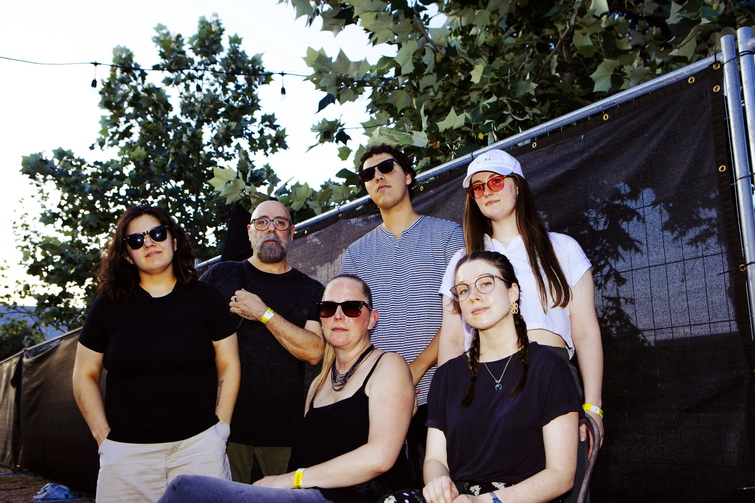A group of six people outdoors at a festival or gathering, posing for a photo in front of a black fence and green trees, with some wearing sunglasses and casual clothing.