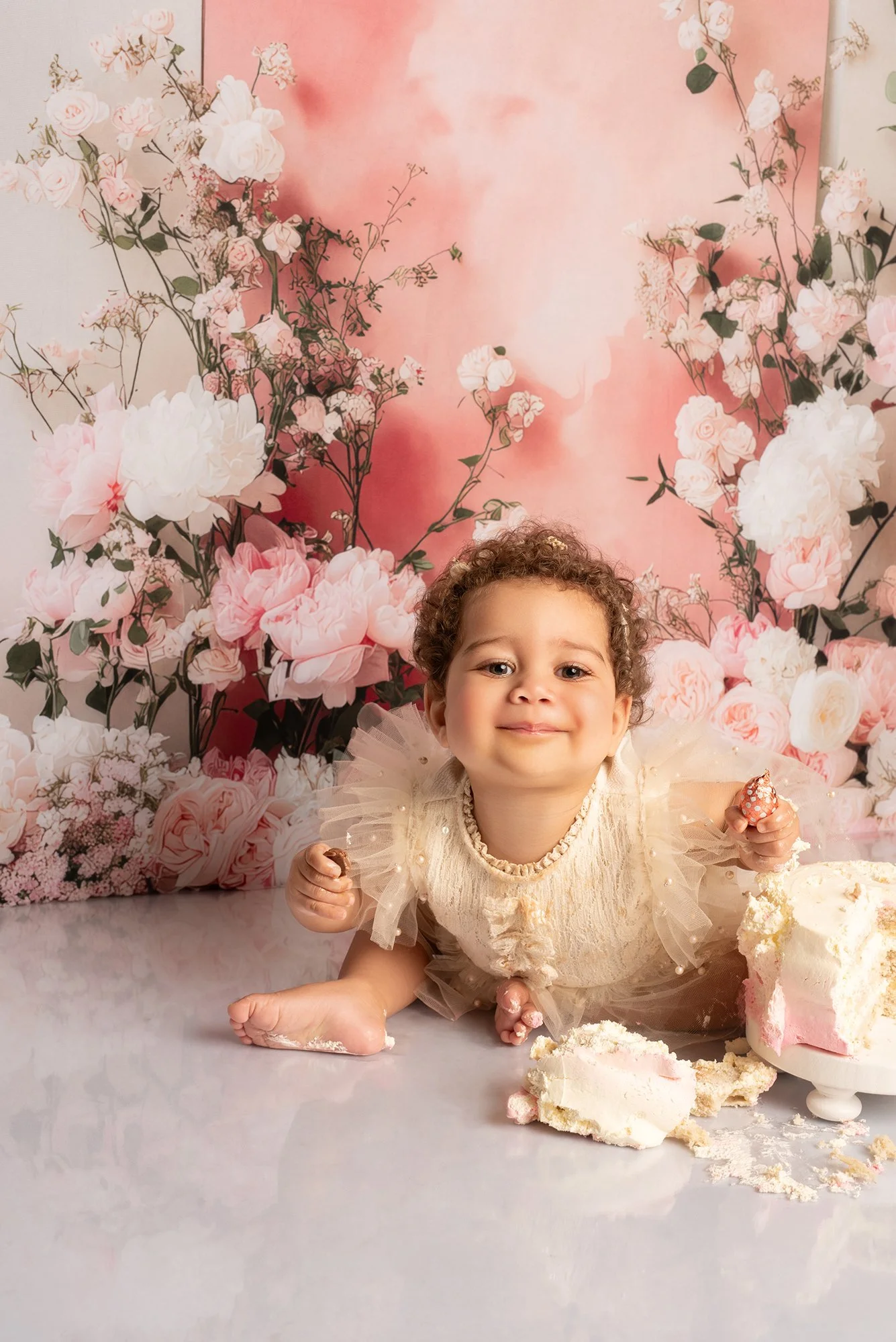 toddler smiling wearing ivory romper with smashed cake infront go her with floral background