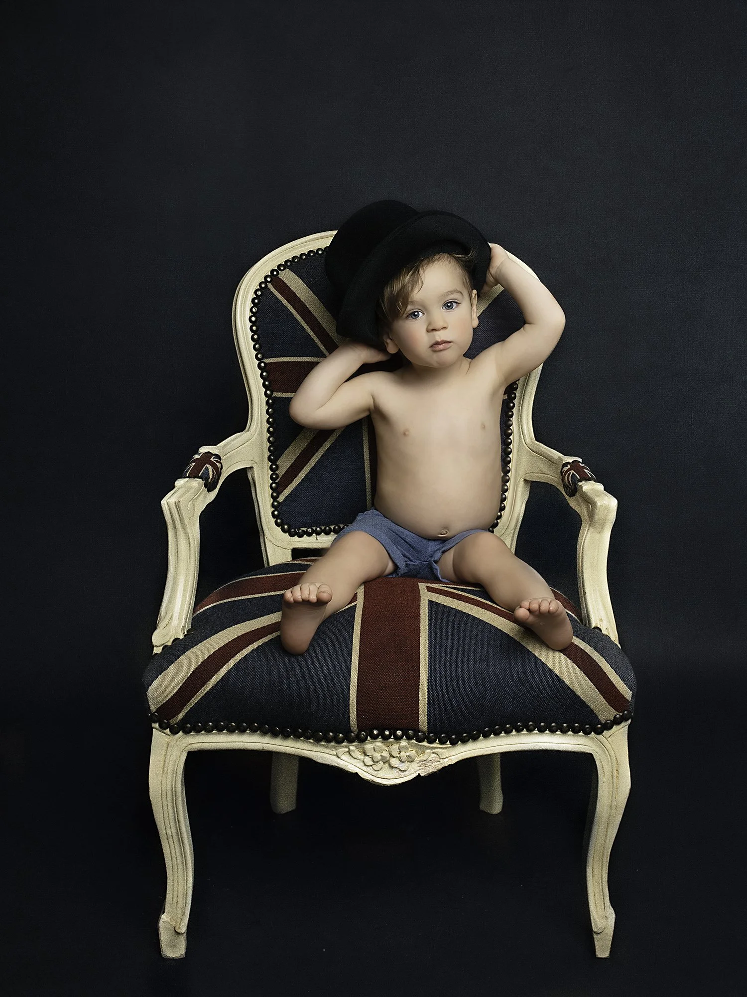 toddler with a hat on a head sitting on a British flag print armchair  with dark navy blue background 