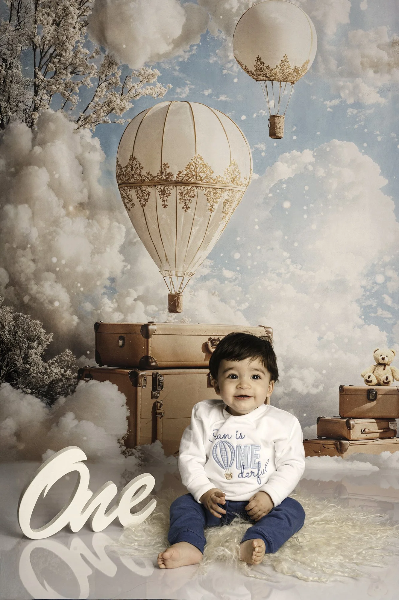 boy in a blue trousers and white top sitting against hot air balloon theme first birthday 