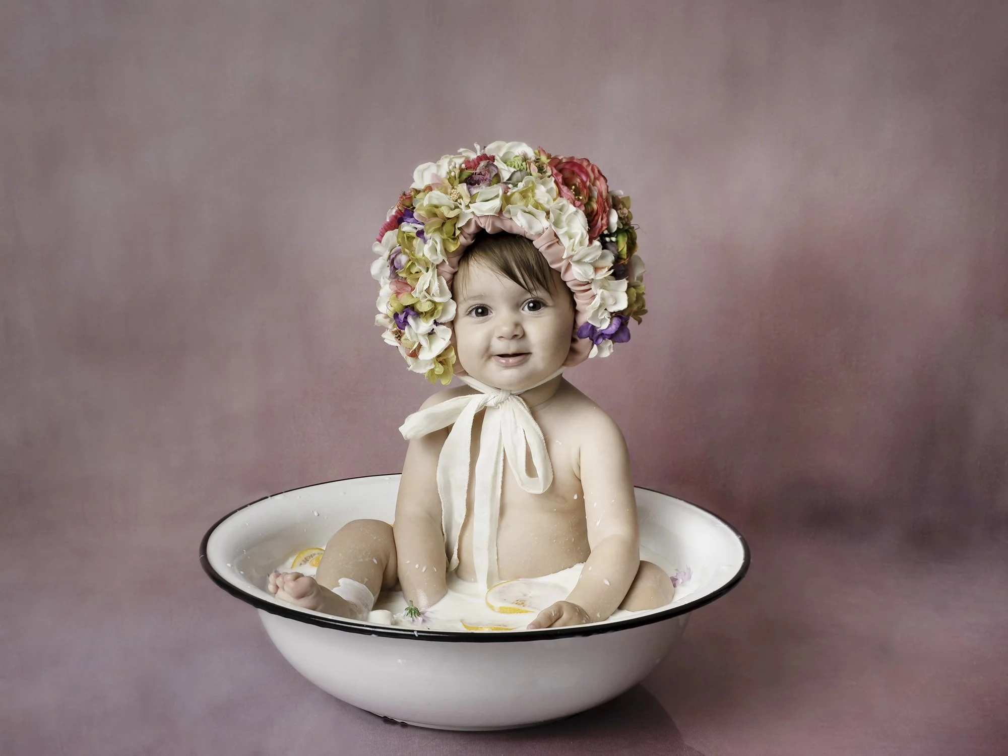 Baby sitting in milk bath filled with flowers and oranges  wearing  floral bonnet