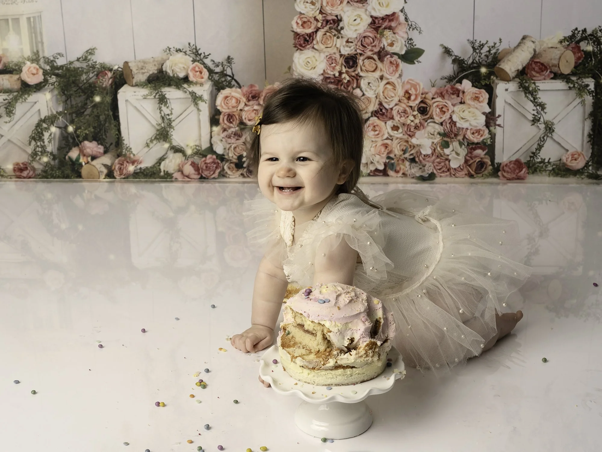 one year old girl in a ivory romper crawling next to cake partially smashed with floral background