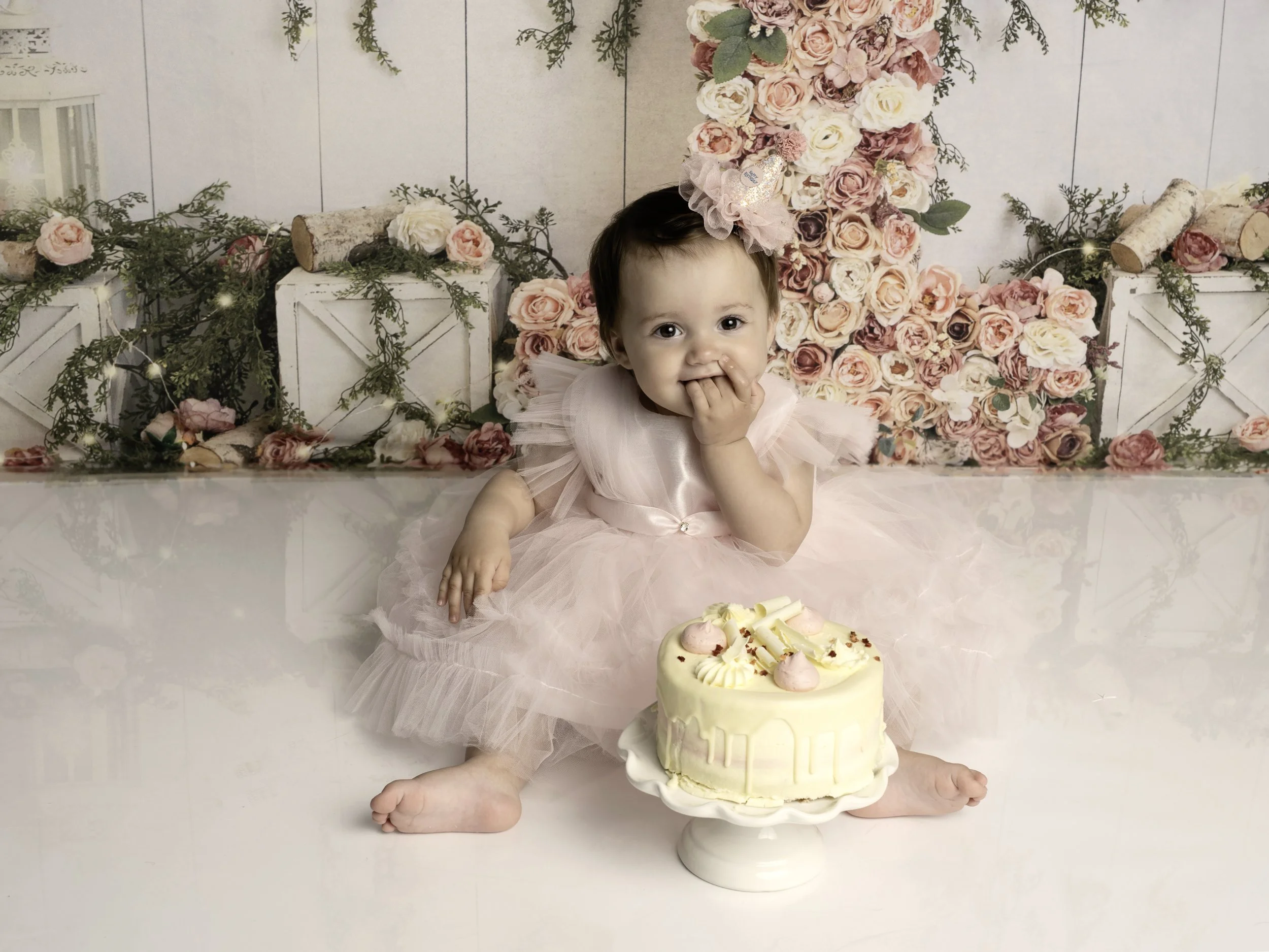 one year old girl in a pink puffy dress with a cake infront of her and floral background 