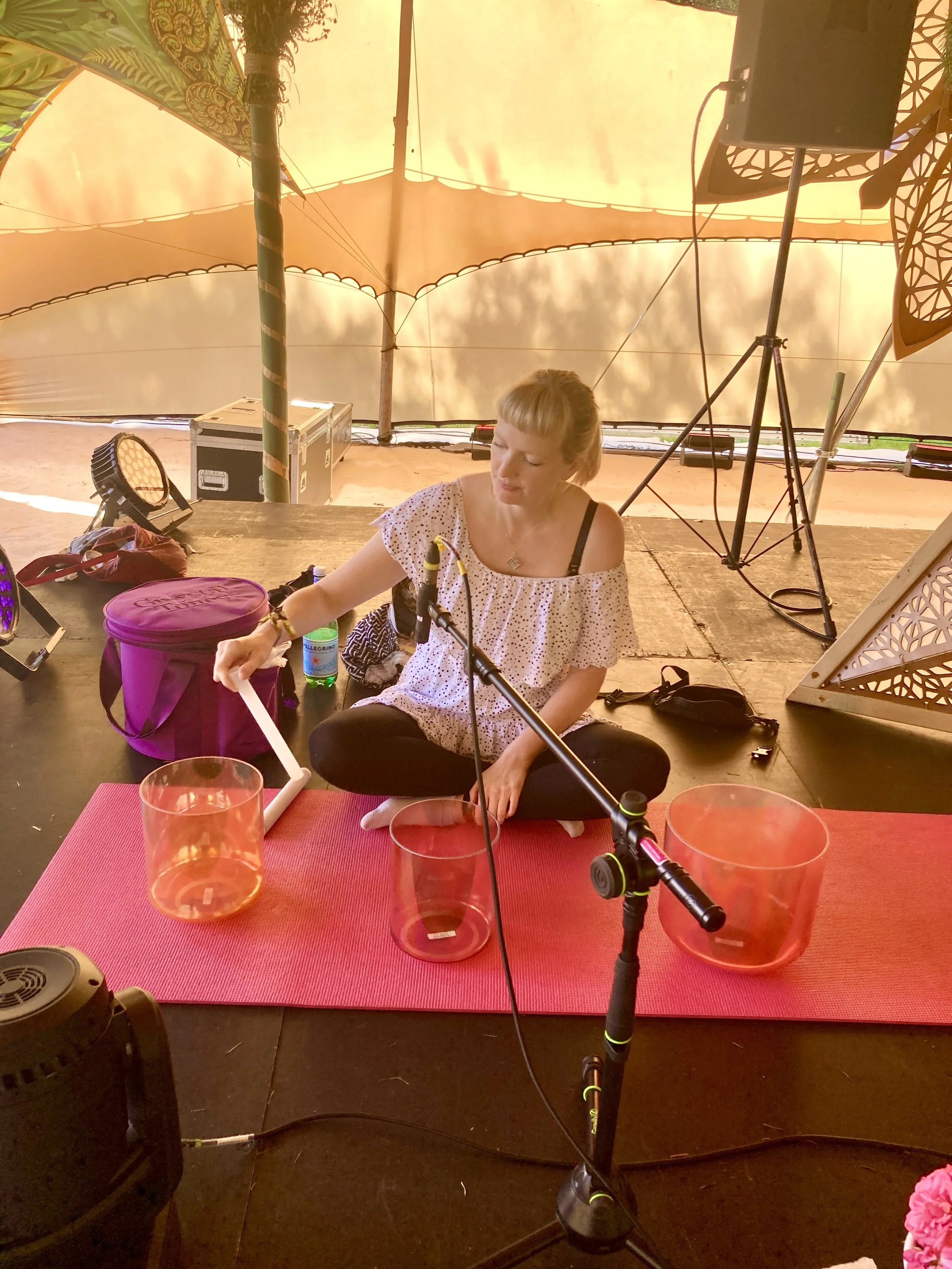 A woman seated on the ground playing crystal singing bowls during a musical performance or meditation session in a tent / outdoor setting at Medicine Festival.