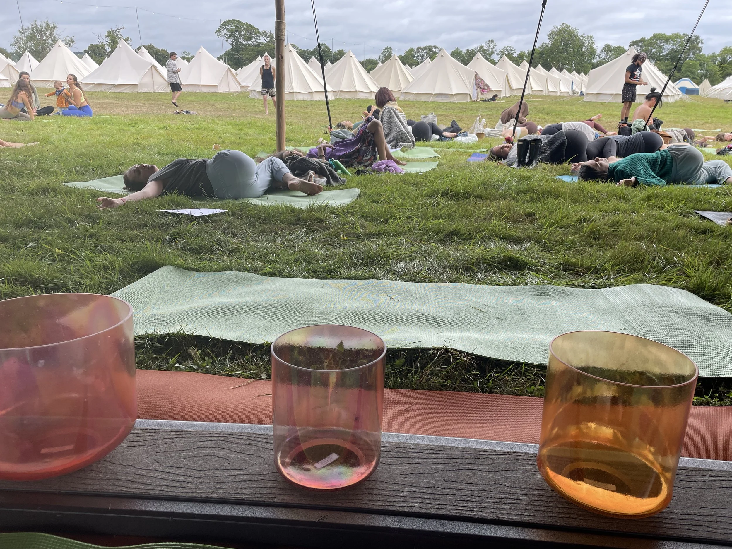 People participating in an outdoor yoga session on a grassy field with tents in the background, viewed from a table with beautiful crystal singing bowls in the foreground.