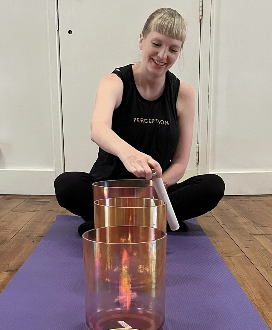 A woman sitting cross-legged on a purple yoga mat, playing crystal singing bowls.