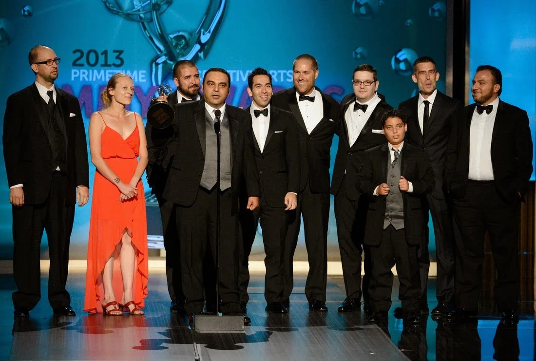 Group of people on stage at an awards ceremony, some wearing tuxedos and one woman in a red dress, with a large screen behind them displaying the year 2013 and the words "Prime Time Emmy Awards."