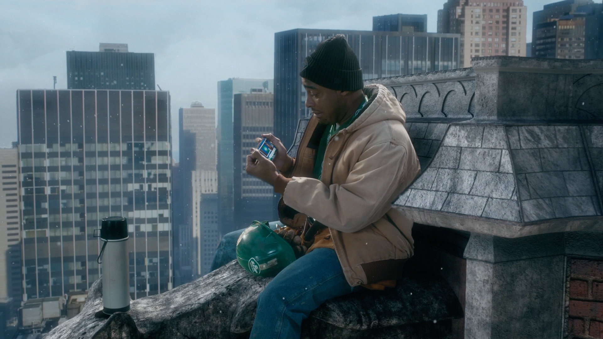 A man in a tan jacket and black beanie sitting on a rooftop ledge in New York City, looking at his phone, with city skyscrapers in the background.