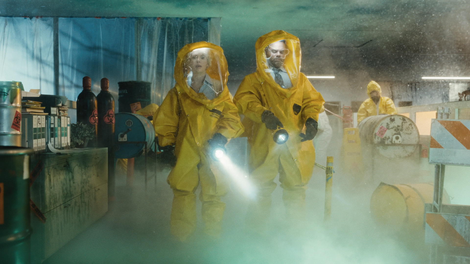 Two people in yellow hazmat suits with face shields, holding flashlights, inside a room filled with fog or smoke, surrounded by hazardous materials and supplies.