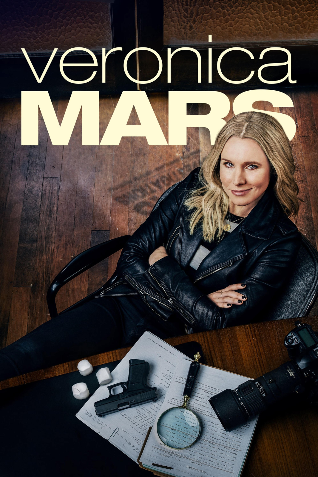 A woman with blonde hair sitting at a table with police equipment, a camera, and documents, looking up at the camera, with the name 'Veronica Mars' displayed above her.