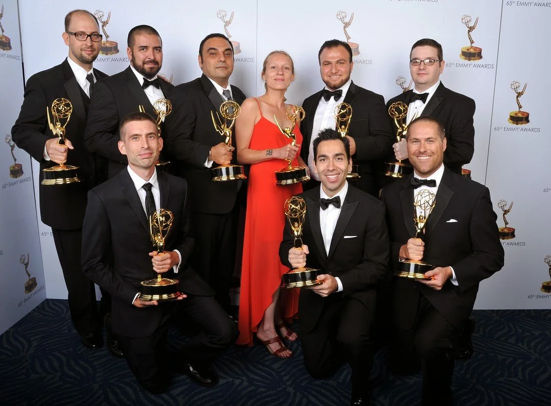 Group of nine people in formal attire holding Emmy awards, posing for a photo at the Emmy Awards event.