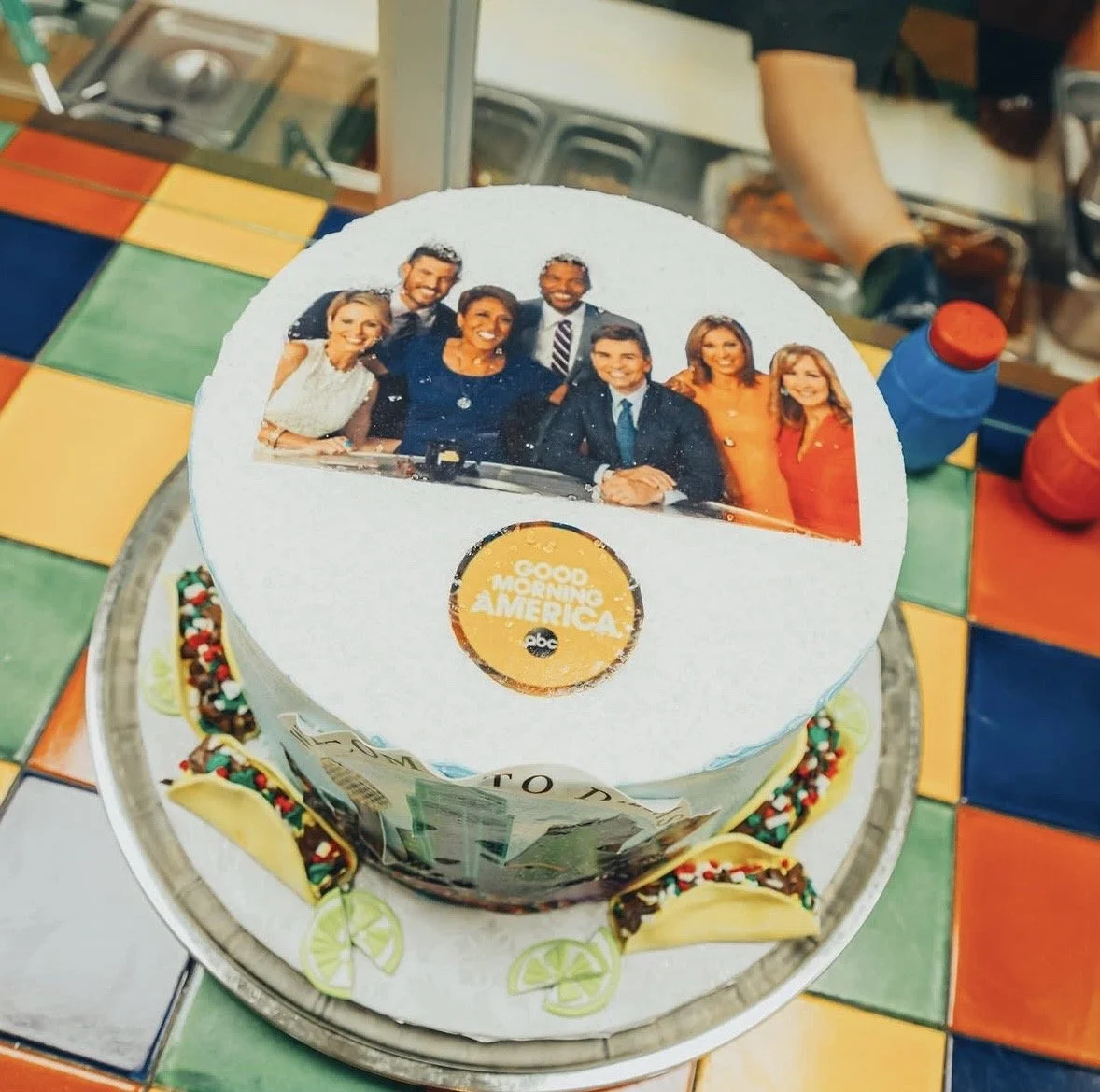 A cake with a photograph of news anchors on top, featuring the 'Good Morning America' logo, placed on a colorful tiled surface.
