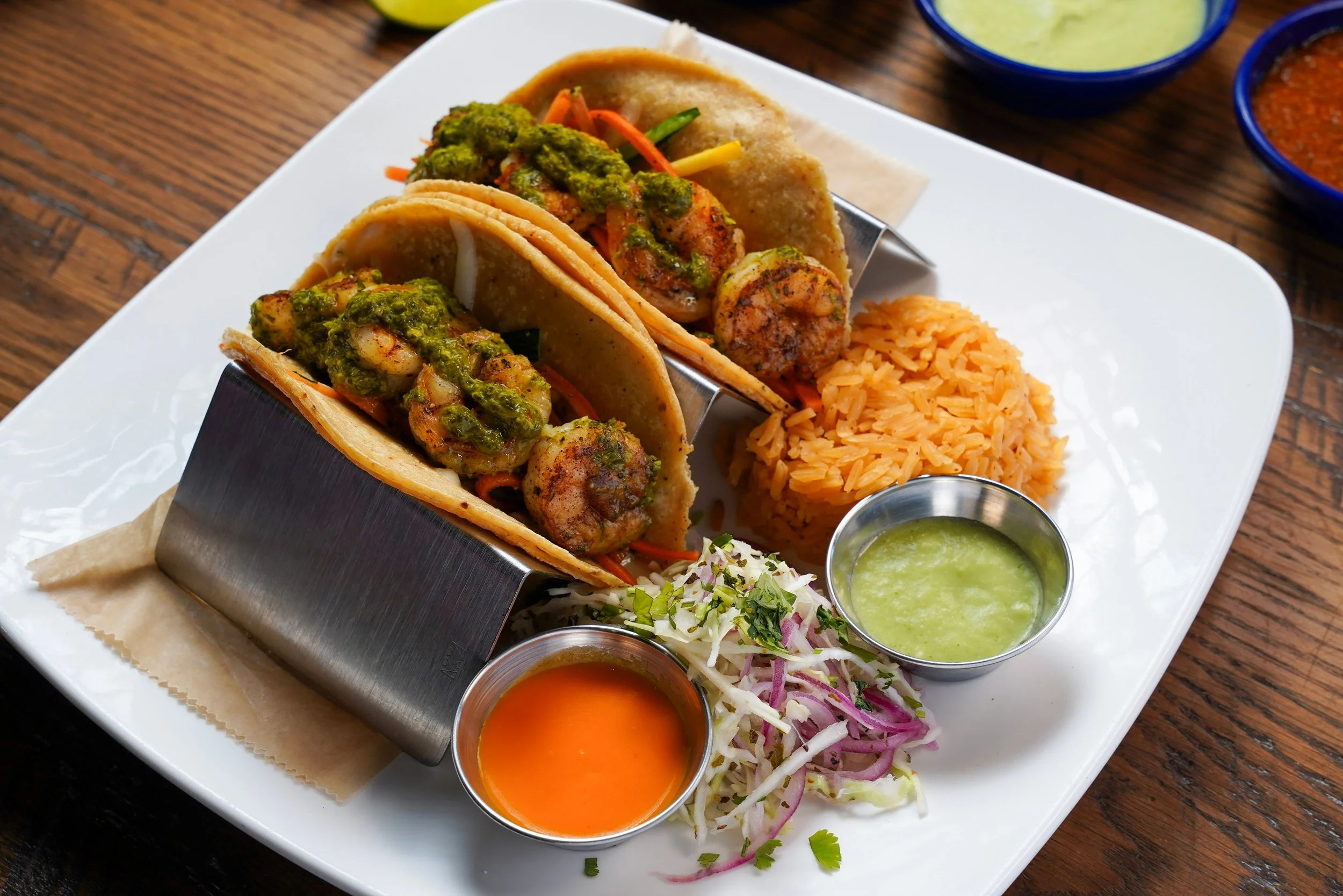 Plate of three soft shell tacos filled with shrimp, topped with green sauce, served with rice, coleslaw, and various sauces on the side, on a white plate on a wooden table.