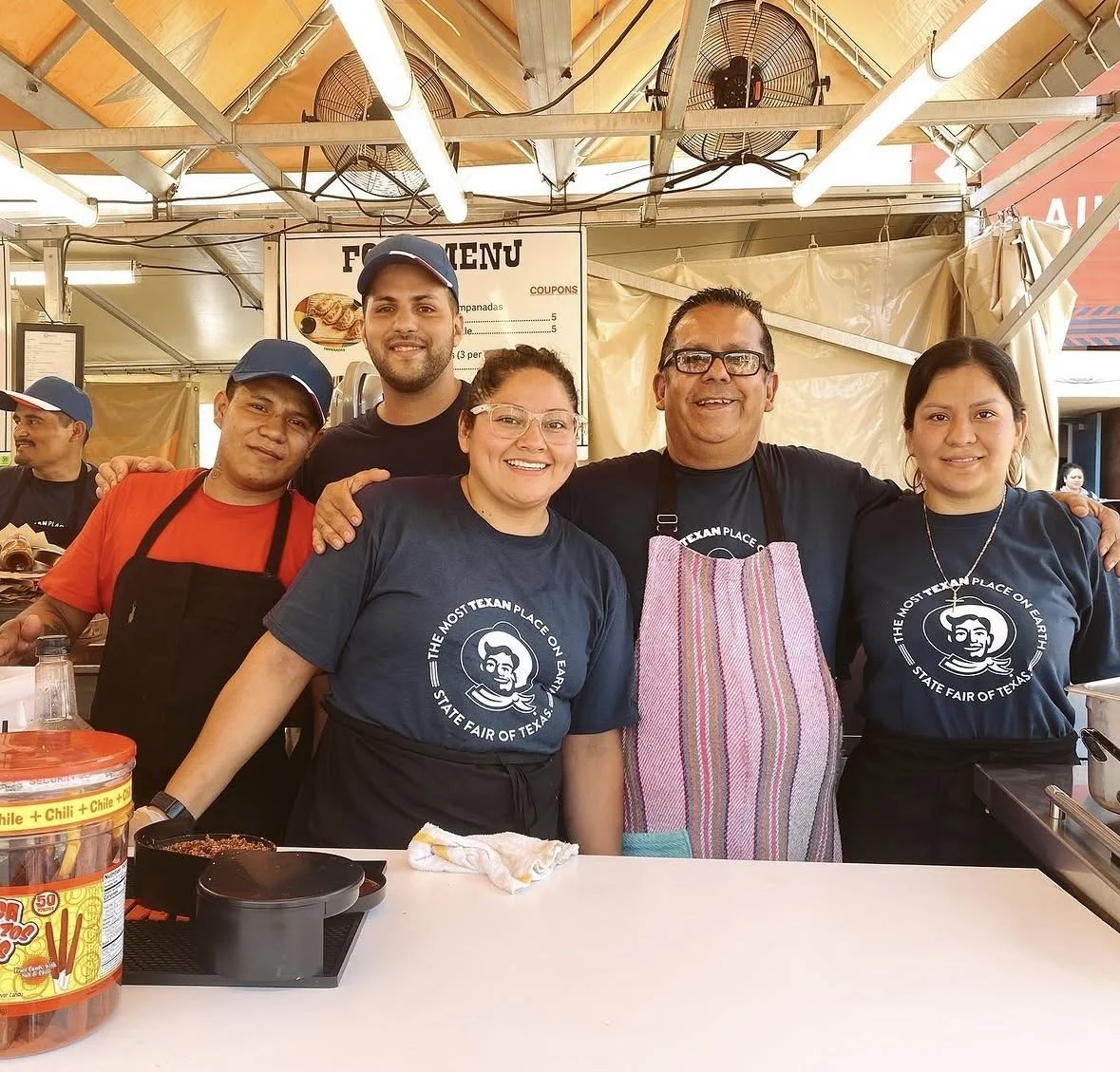Group of five people standing behind a food stall at the Texas State Fair of Texas, wearing black T-shirts with a mascot logo and an apron.