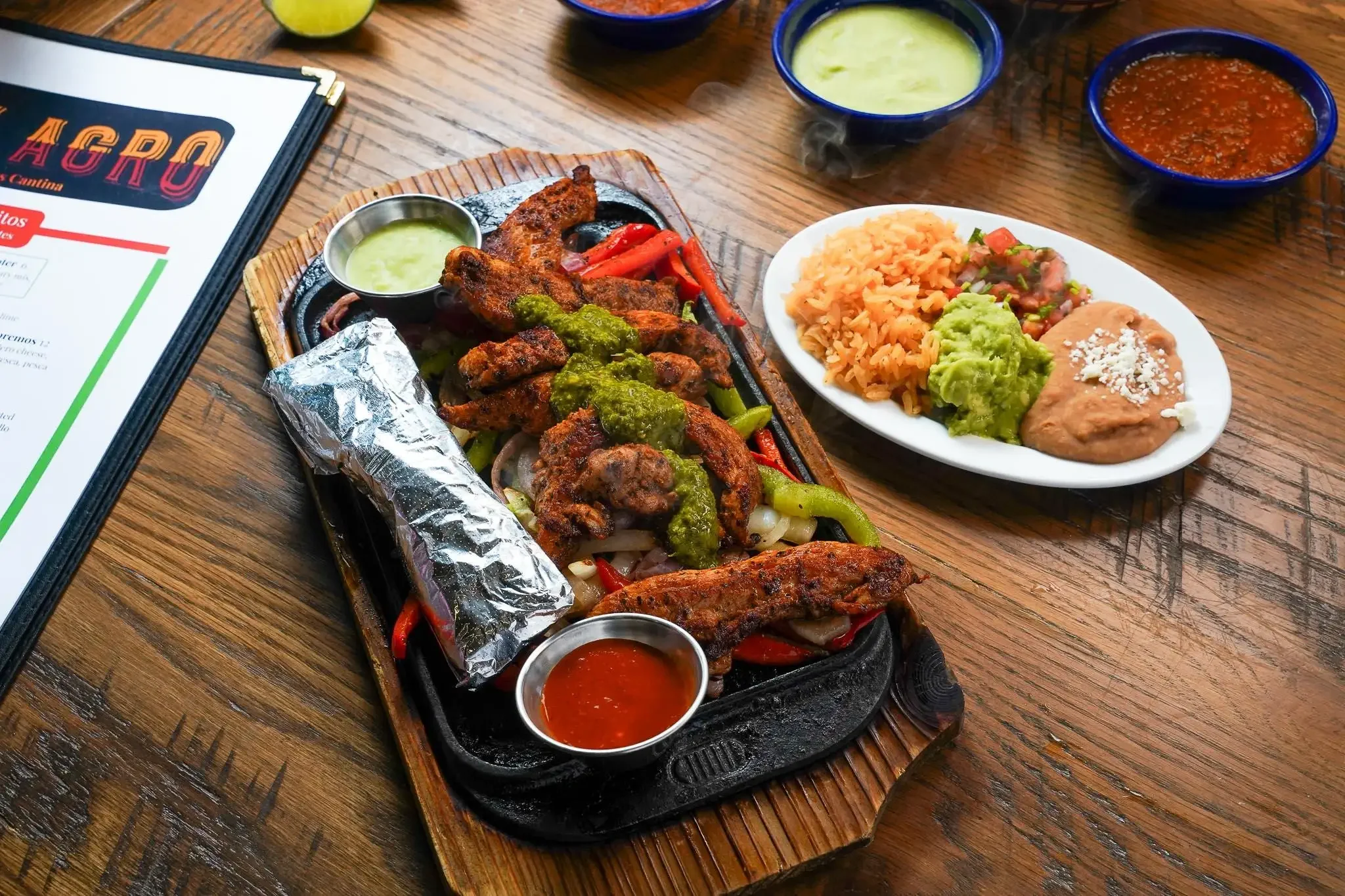 A wooden table with a Mexican meal including grilled meat with green sauce, grilled vegetables, rice, refried beans with cheese, and various salsas in small bowls.