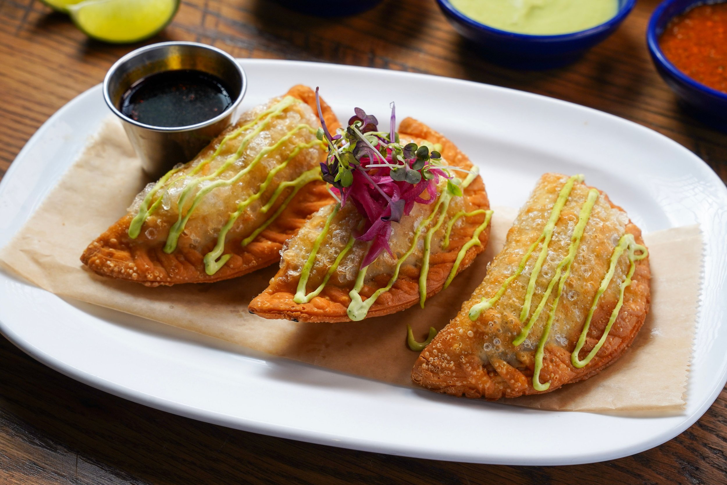 Three fried fish fillets topped with lime and microgreens on a white plate, drizzled with green sauce, accompanied by dipping sauces in small bowls.