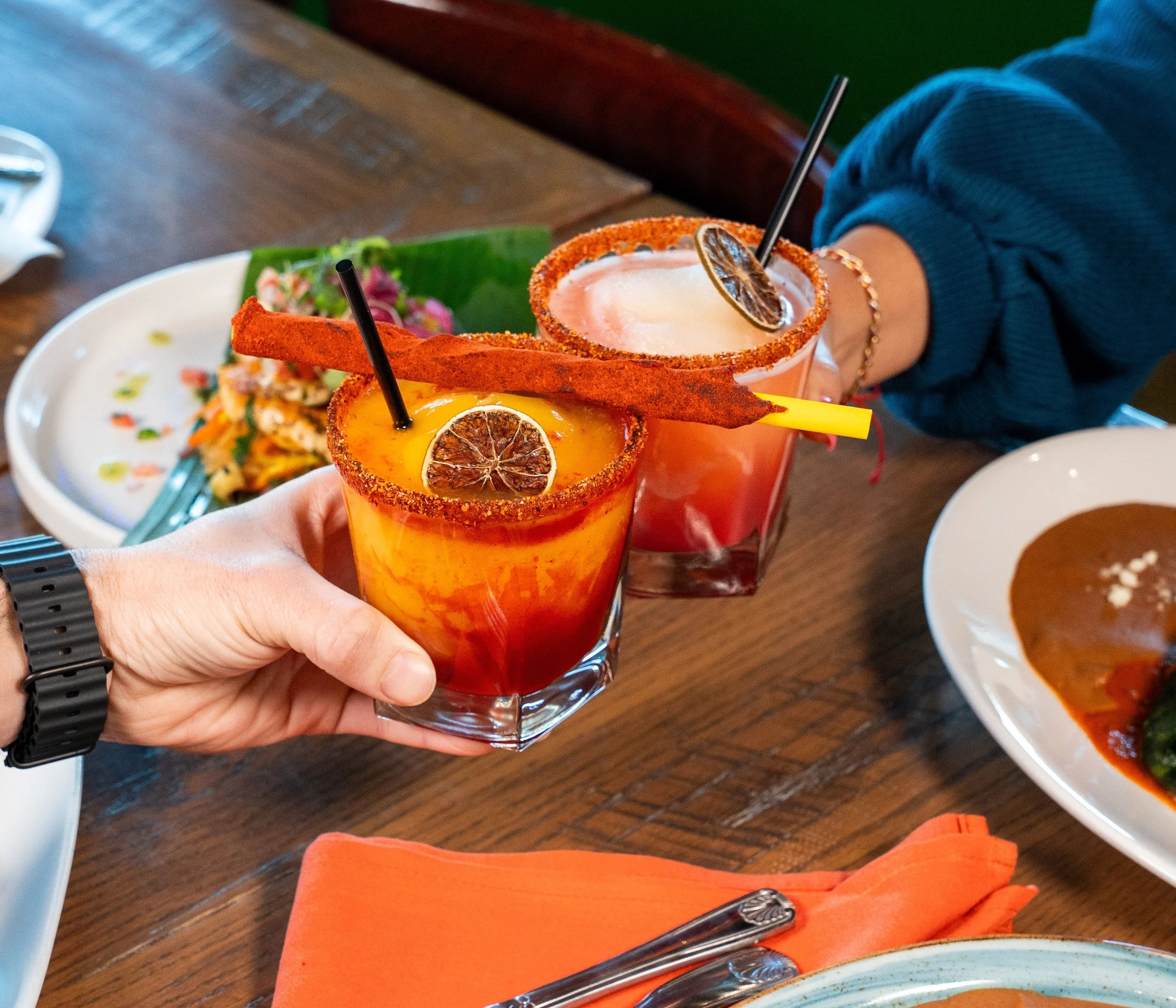 Two colorful cocktails with salted rims, topped with dried lime slices, held by a person wearing a blue sweater, on a wooden table with plates of food.