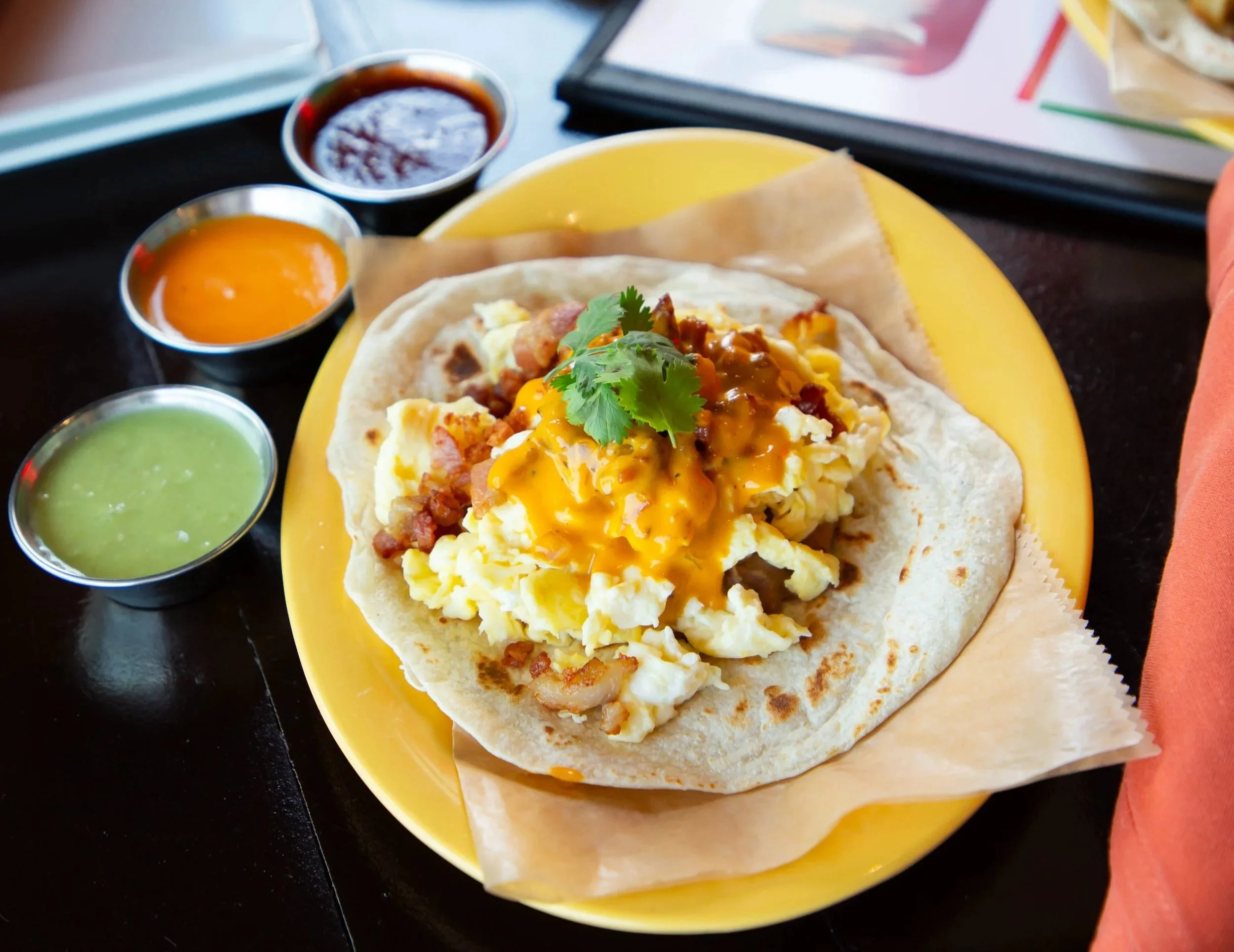 A breakfast tacos dish with scrambled eggs, bacon, cheese, cilantro, and hot sauce, served on a corn tortilla on a yellow plate with small cups of green and red salsa on the side.