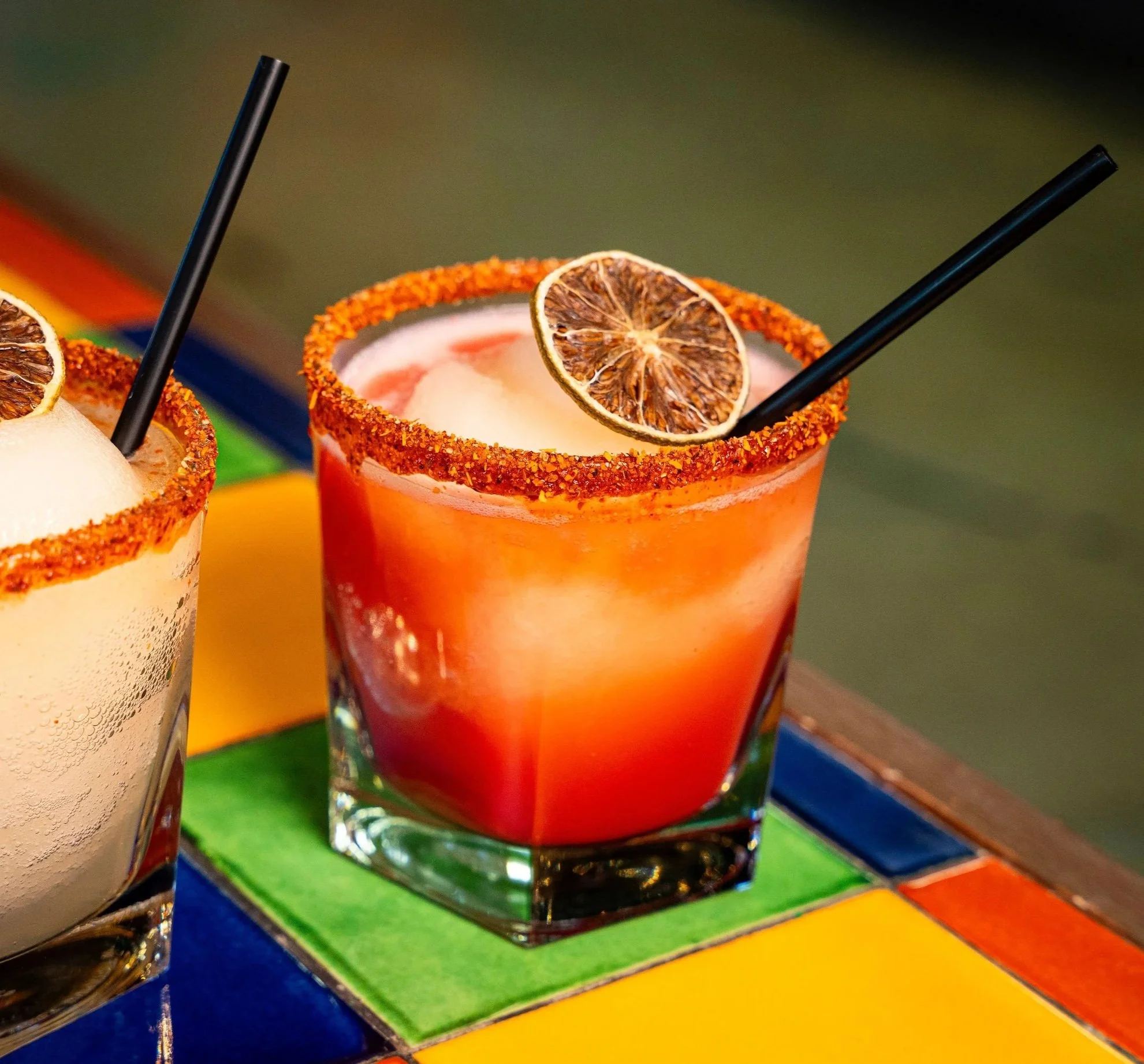 Close-up of a cocktail glass with a chili salt rim, garnished with a dried lime slice and a black straw, on a colorful striped table.