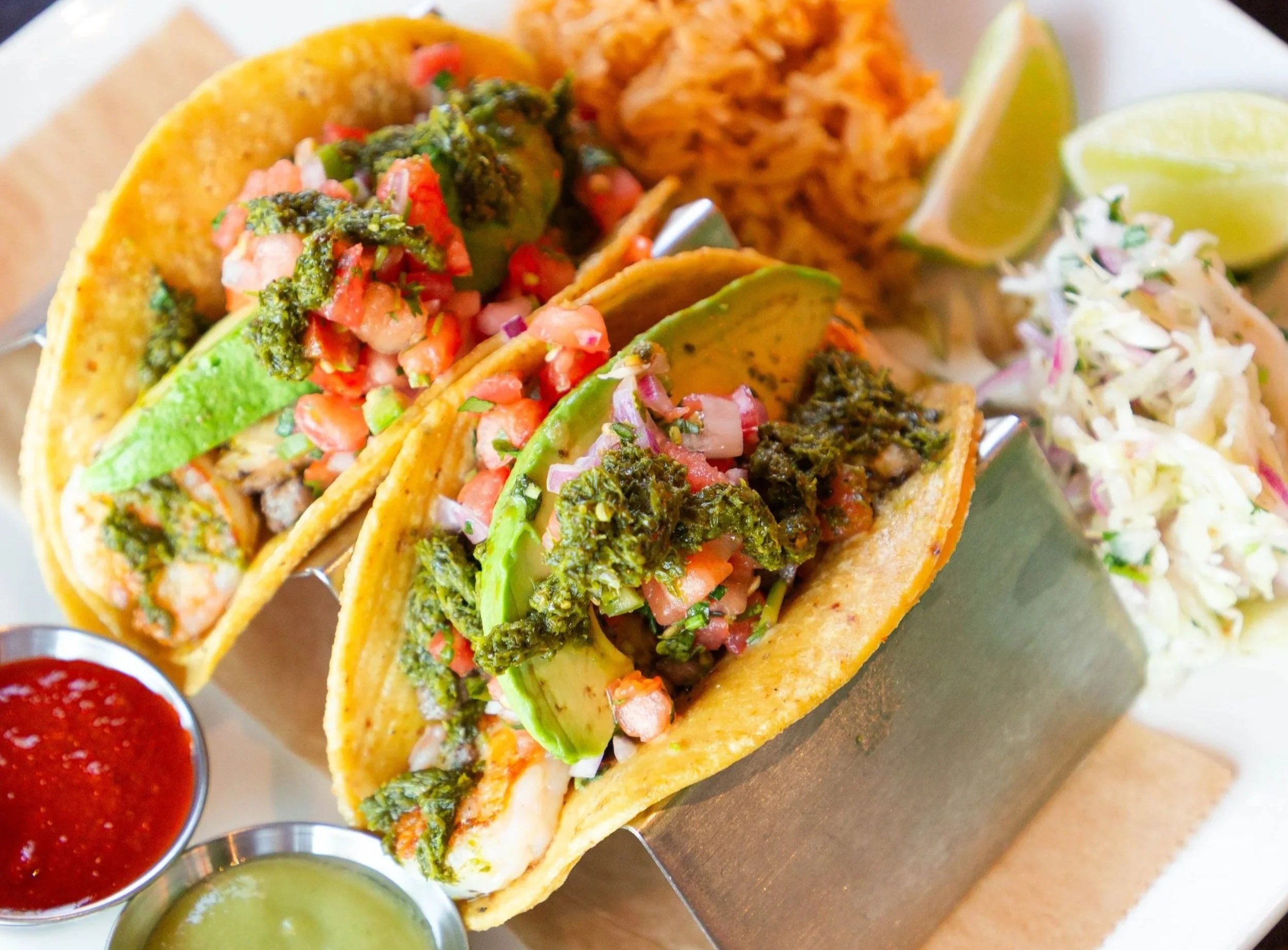 Close-up of two tacos filled with chopped vegetables and sauce, served with lime wedges and three small bowls of salsa and green sauce on a tray.