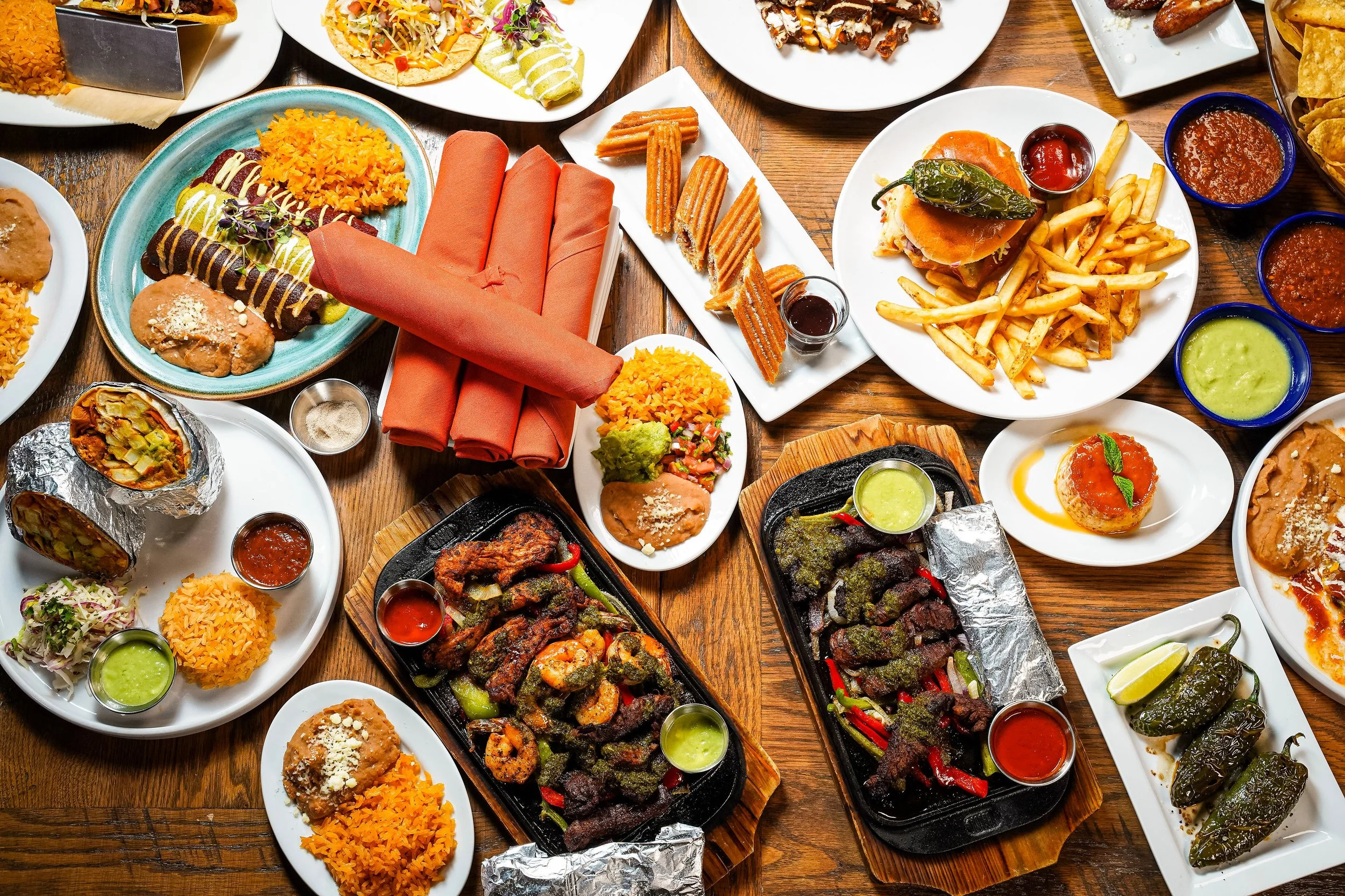 A table filled with various Mexican dishes including rice, grilled meats, tacos, guacamole, salsa, and drinks.