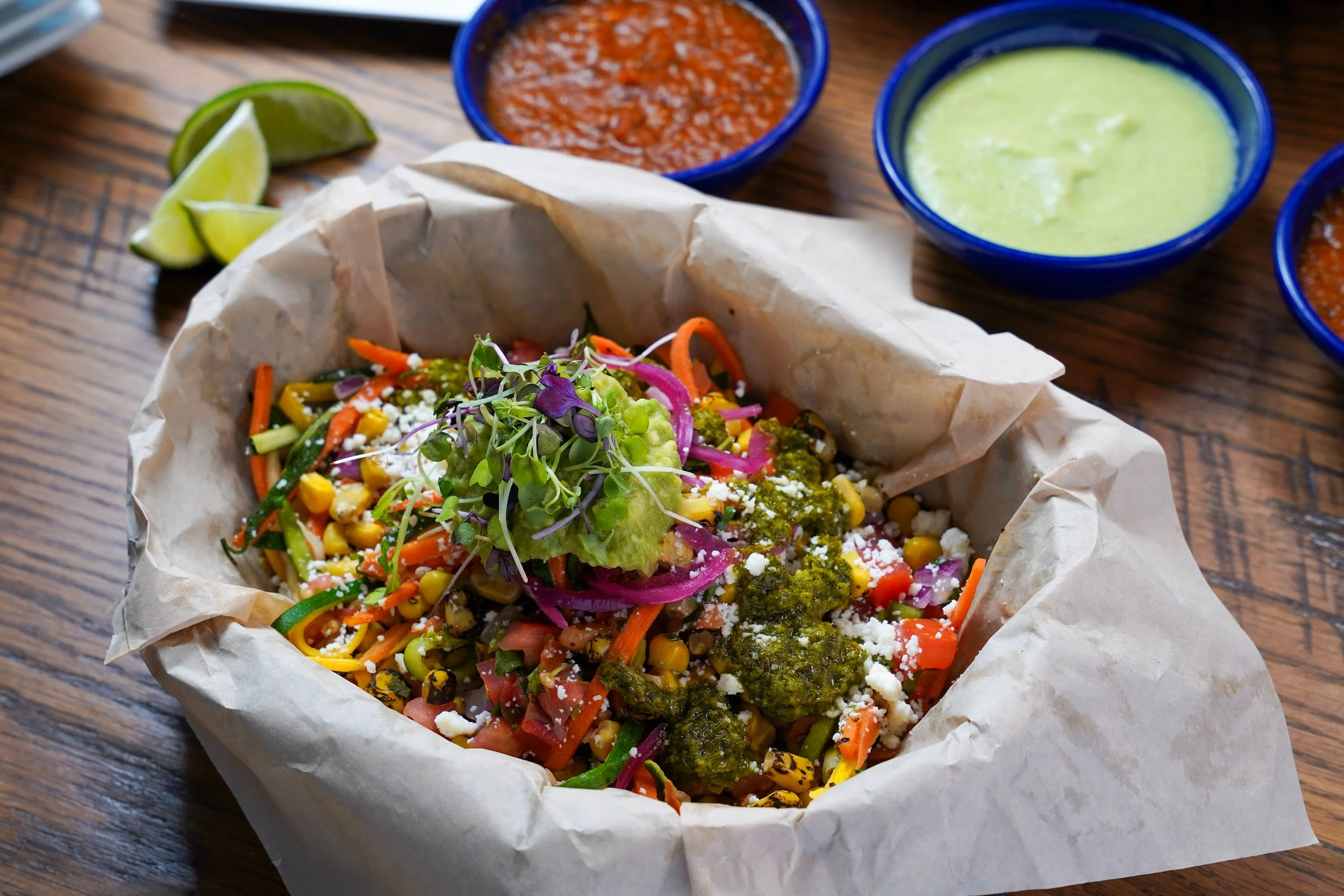 Colorful vegan bowl filled with mixed vegetables, topped with microgreens, served in a paper-lined basket on a wooden table with lime wedges and two small bowls of salsa and guacamole in the background.