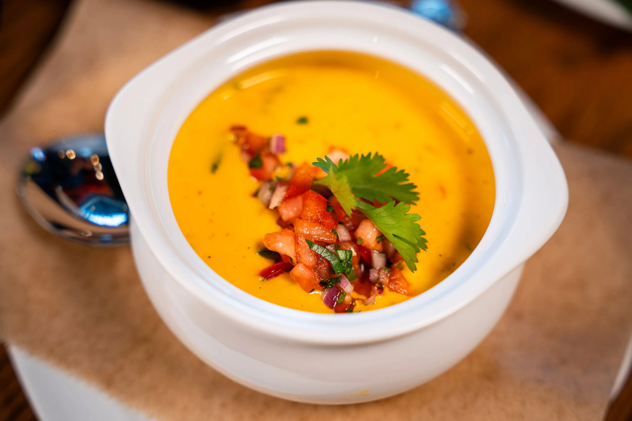 A white bowl of creamy yellow soup garnished with chopped tomatoes, cilantro, and red onion, placed on a brown surface.