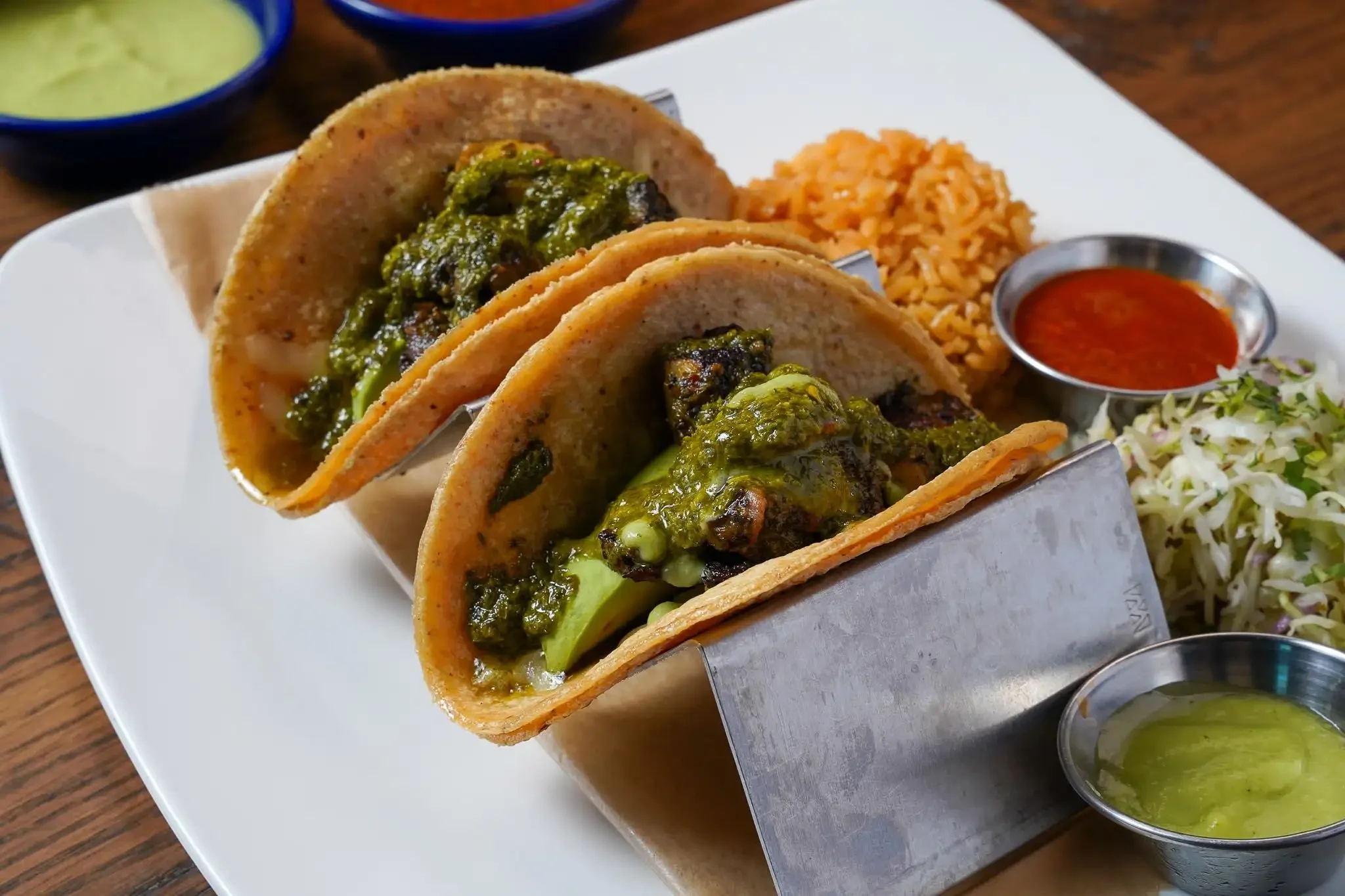 Three tacos with meat, green salsa, and avocado served on a white plate with rice, beans, and salsa on the side.