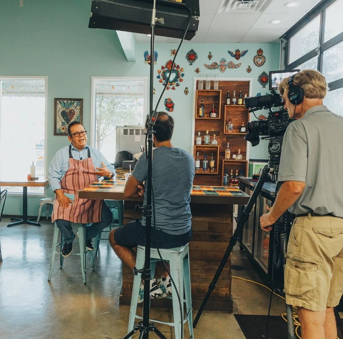 Filming of a television interview or documentary inside a colorful cafe with artwork on the walls and various bottles on a wooden shelf. A man with glasses, in a blue shirt and apron, is speaking into a microphone while two camera operators record him.