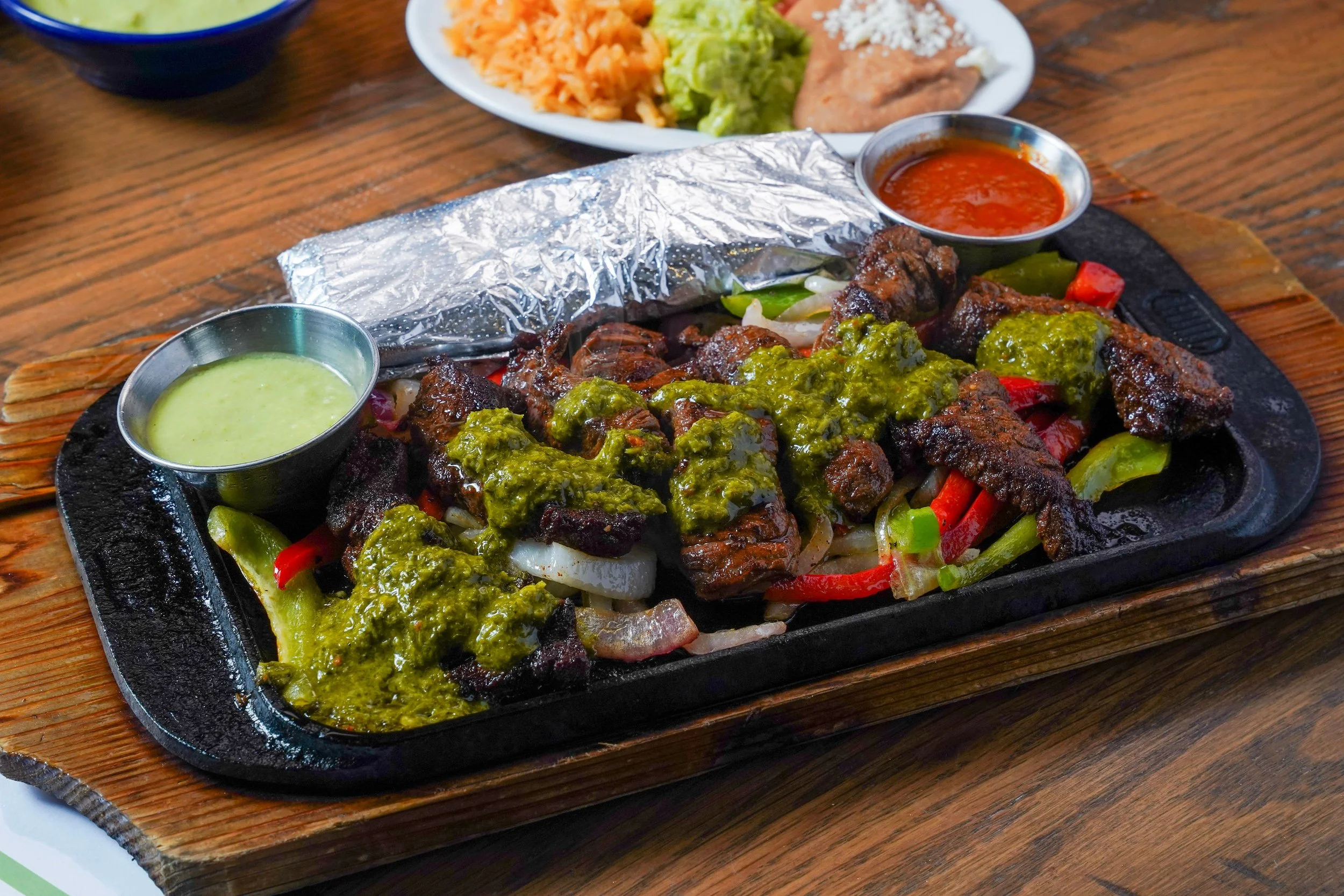 Fajitas with grilled meat and vegetables, topped with green sauce on a sizzling platter, served with side dips of green sauce and red salsa, with rice, guacamole, and refried beans in the background.