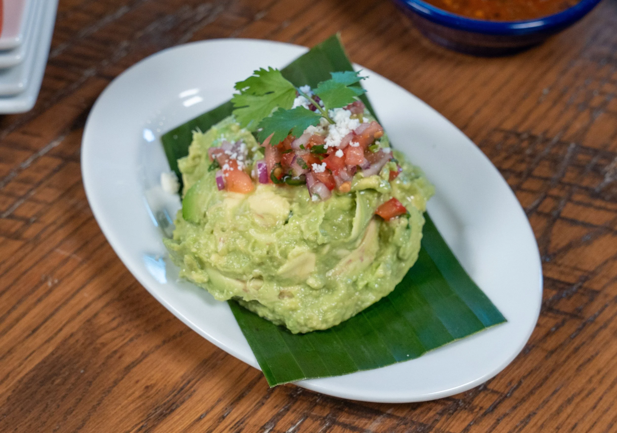 A serving of guacamole topped with chopped tomatoes, onions, and cilantro, garnished with a sprig of cilantro, served on a white plate with a banana leaf and a green banana leaf underneath, on a wooden table.