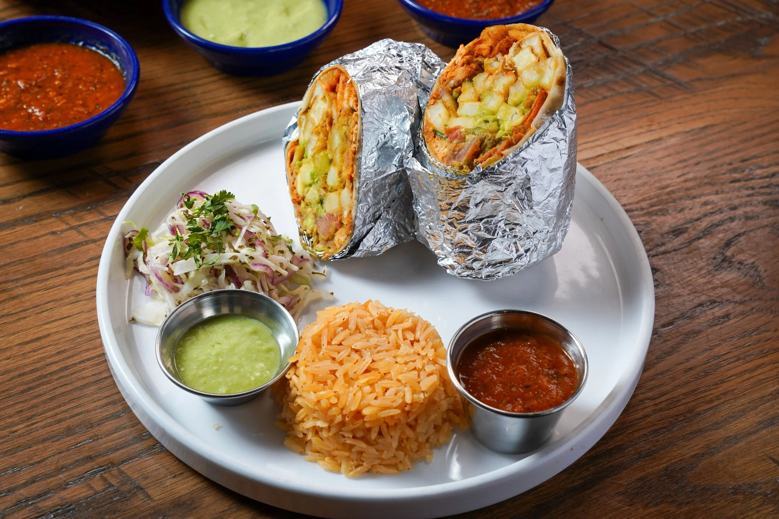 A plate of Mexican food with a burrito cut in half showing inside ingredients, served with side servings of rice, coleslaw, and various salsas and sauces on a round white tray on a wooden table.