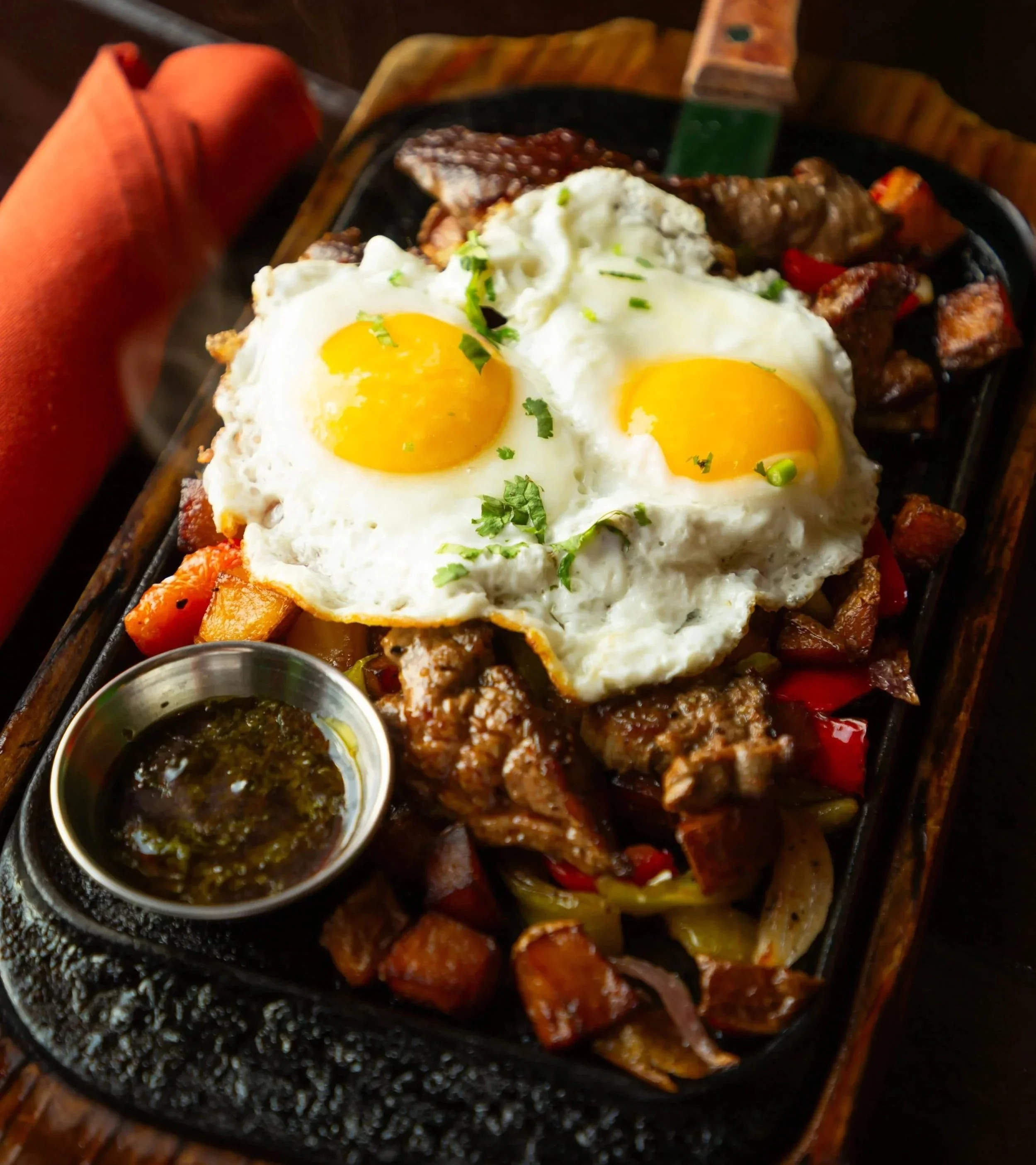 Skillet with sunny side up eggs, cooked beef, grilled vegetables, and green sauce in a small container, served on a black sizzling plate.
