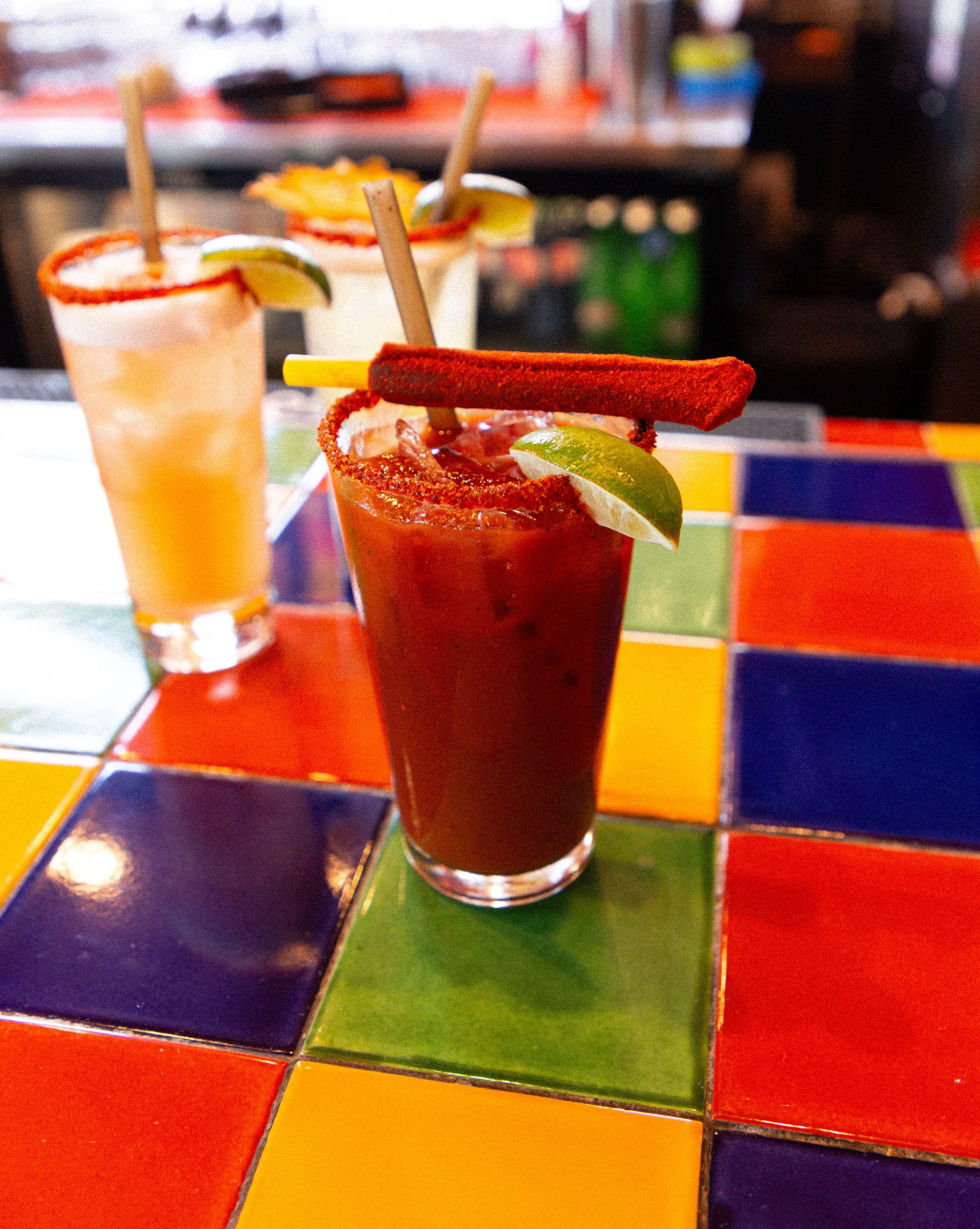 Close-up of two cocktails on a colorful tiled table in a restaurant. One drink is red with lime and a salted rim, garnished with a lime wedge and a chili pepper. The other drink is yellow with a salted rim, also garnished with lime and chili pepper.