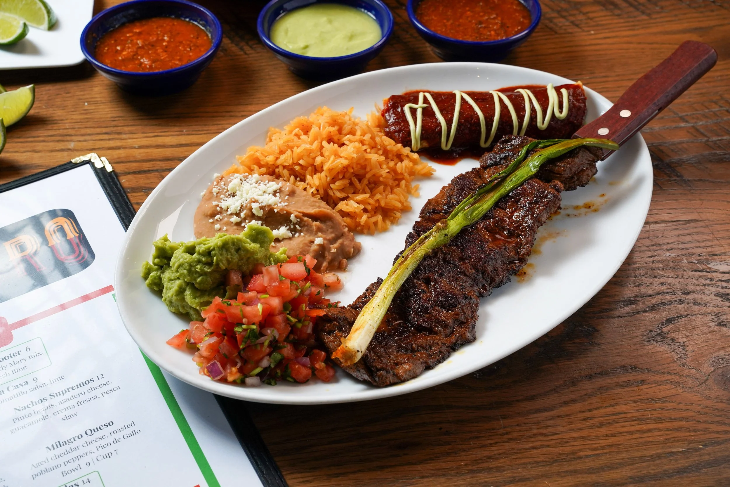 A white plate with Mexican food including rice, refried beans with cheese, a beef steak with green onion, a grilled beef sausage topped with mayonnaise, and a colorful tomato, onion, and cilantro salad. There are three small blue bowls with sauces in the background and lime wedges to the side.