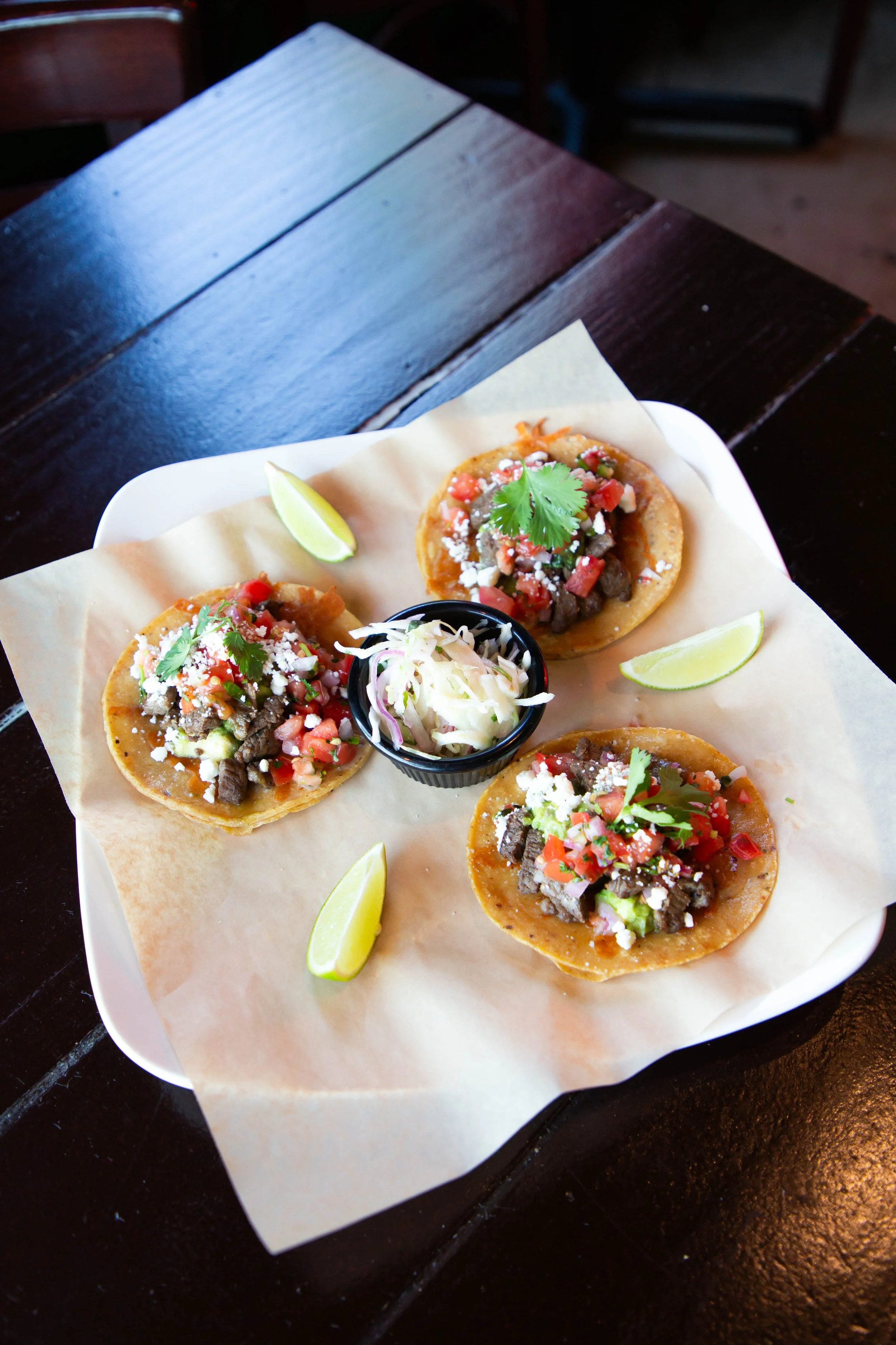 Close-up of a white plate with three tacos topped with diced tomatoes, cilantro, crumbled cheese, and ground meat, garnished with lime wedges and a cup of shredded cabbage on a black table.