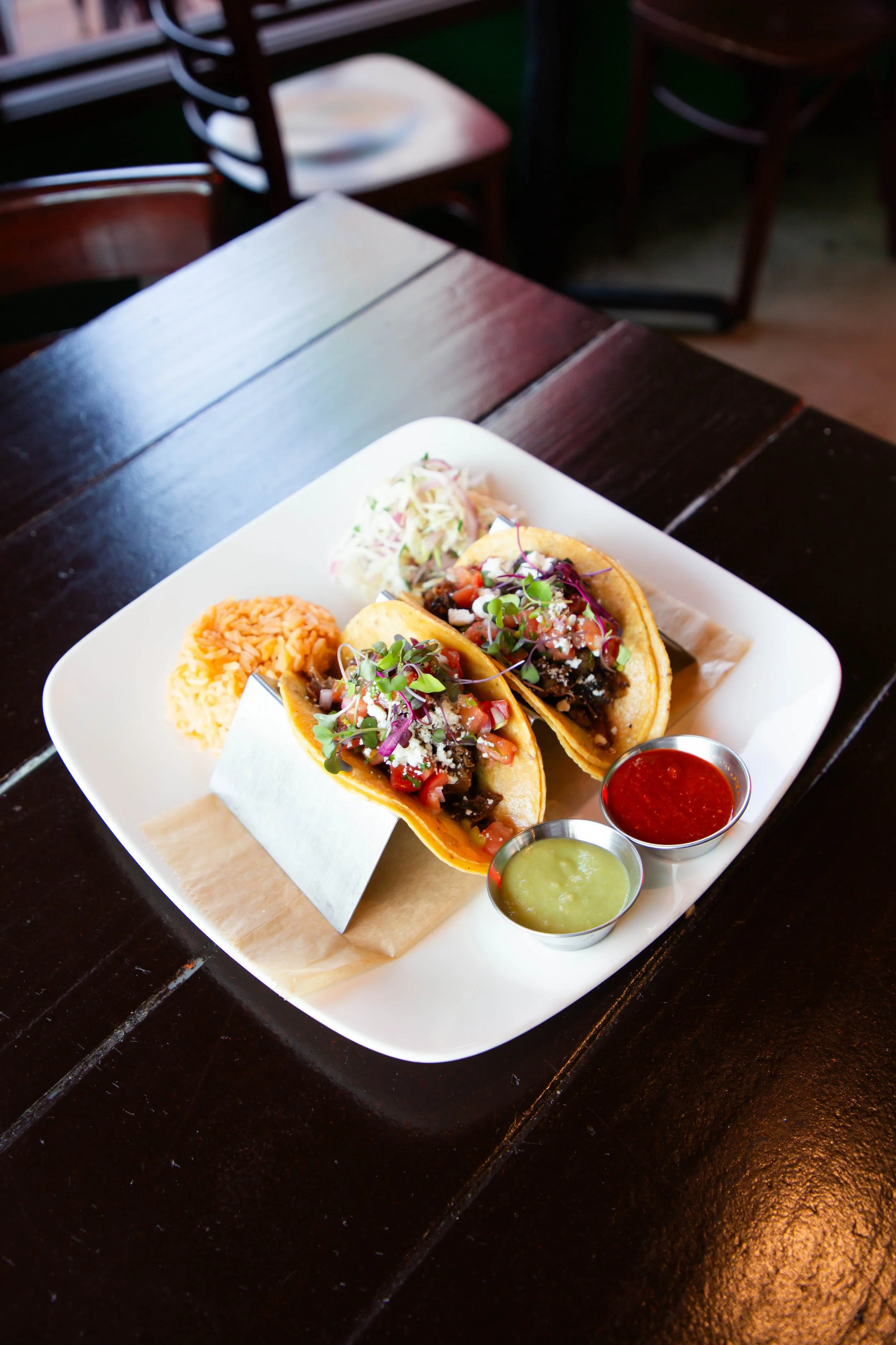 A white plate with two small tacos filled with meat, chopped vegetables, and microgreens, accompanied by rice, coleslaw, and three cups of salsa and green sauce on a dark wooden table.
