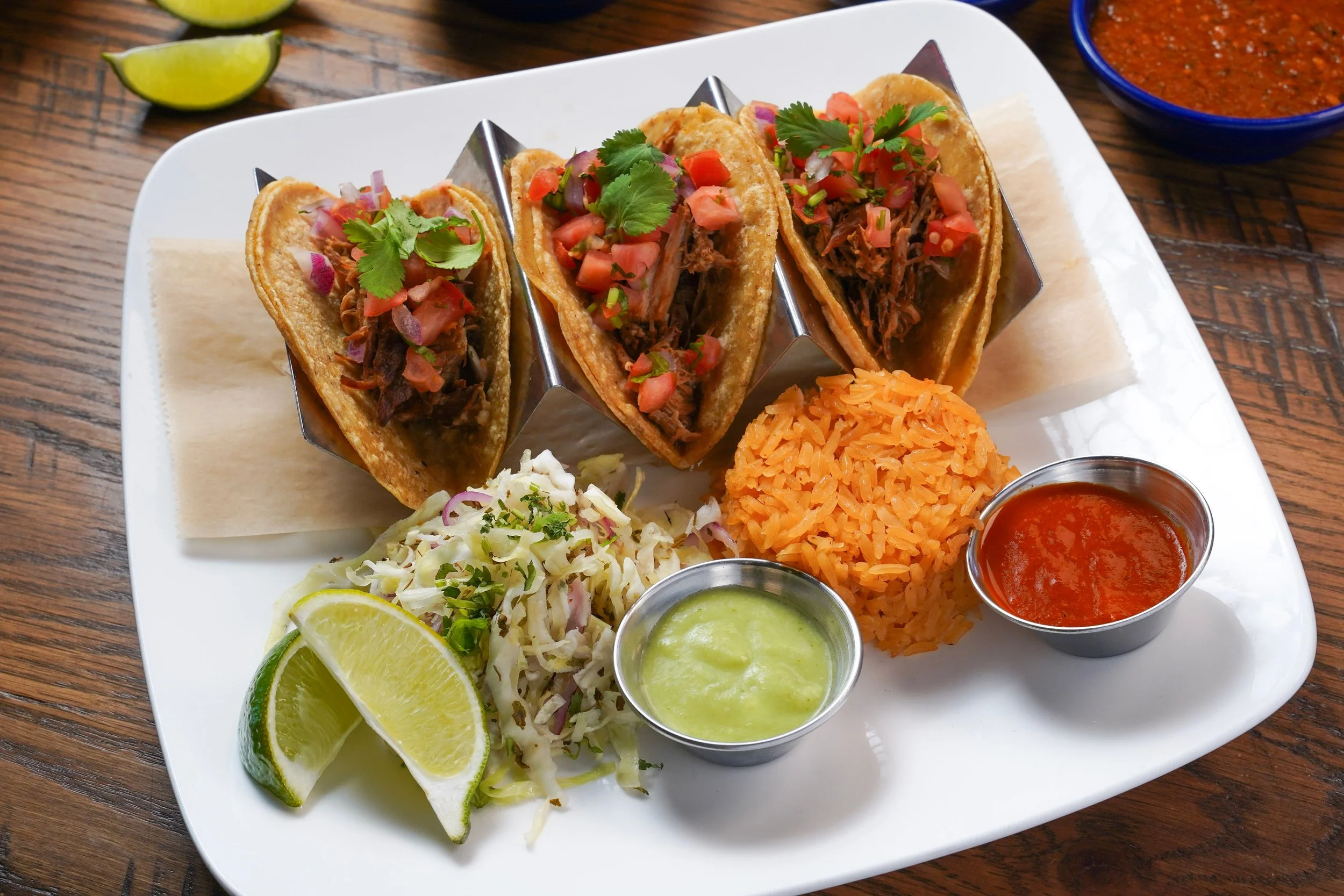 Three street tacos topped with chopped tomatoes, onions, and cilantro, served on a white plate with lime wedges, shredded cabbage, and various sauces including green and red salsa.