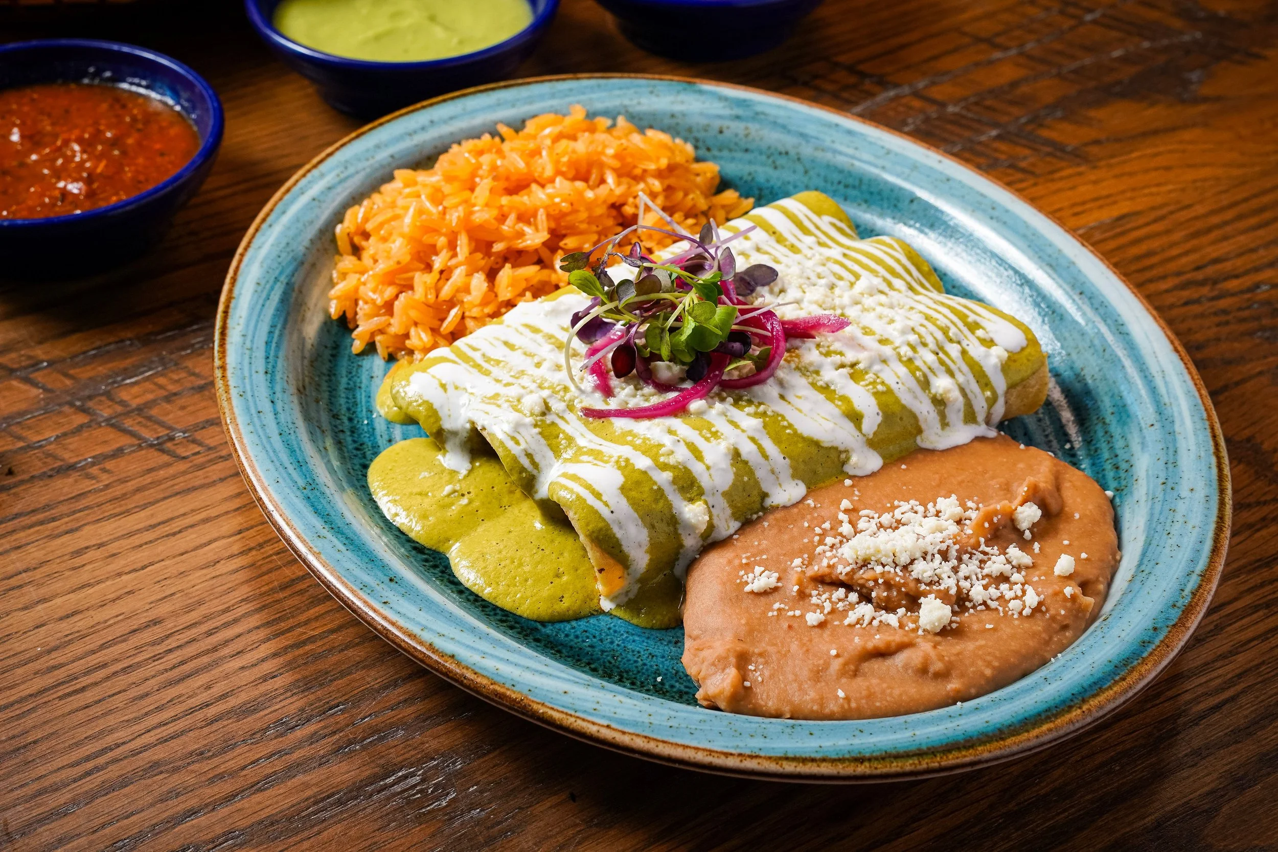 Plate with Mexican food including an enchilada topped with sour cream and pickled red onions, a side of Mexican rice, and various sauces in small bowls on a wooden table.